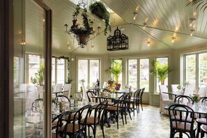 A sunroom with a glass table and chairs, surrounded by large windows and potted plants.