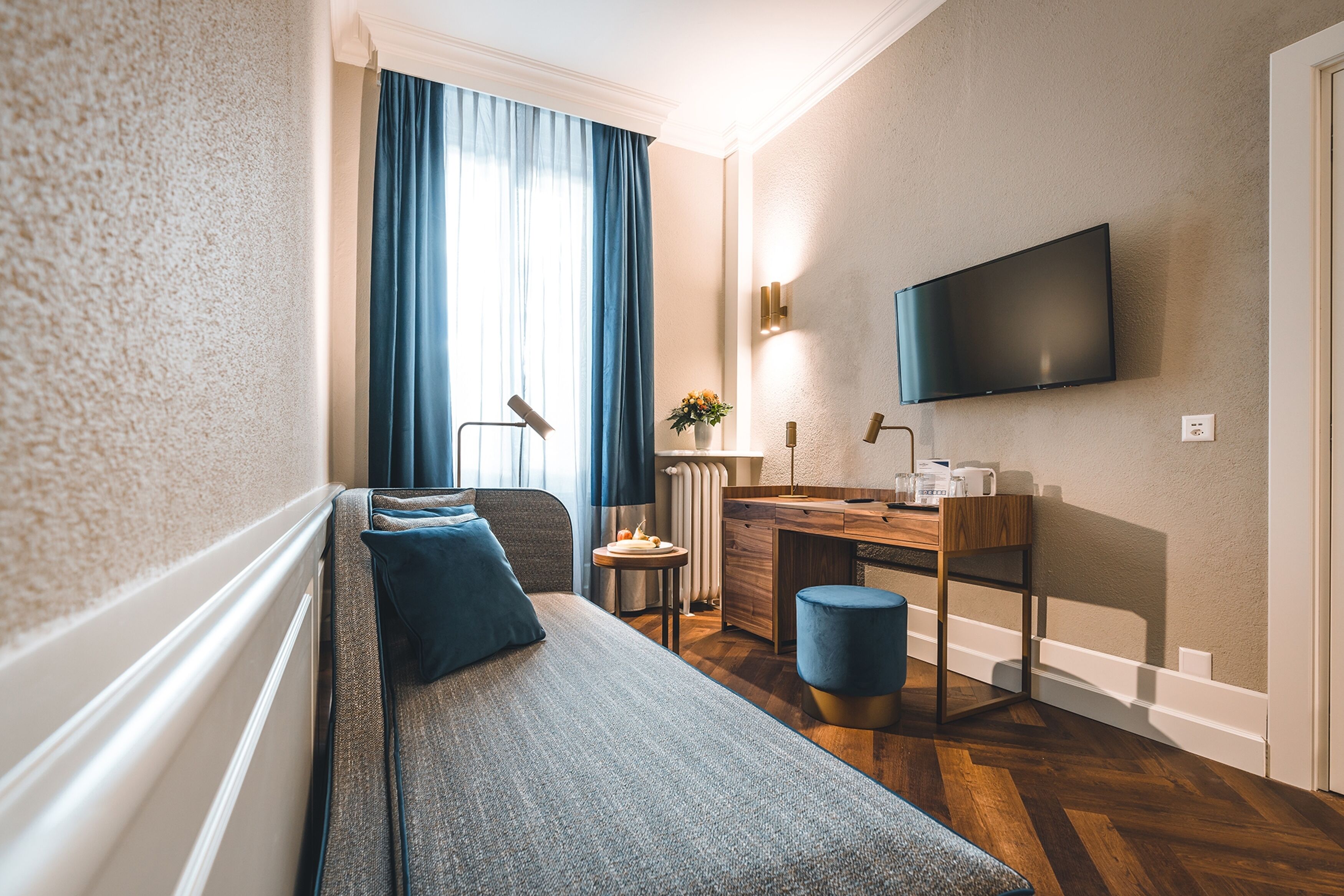 A modern hotel room with a sofa, a small desk, a television, and a window with curtains.