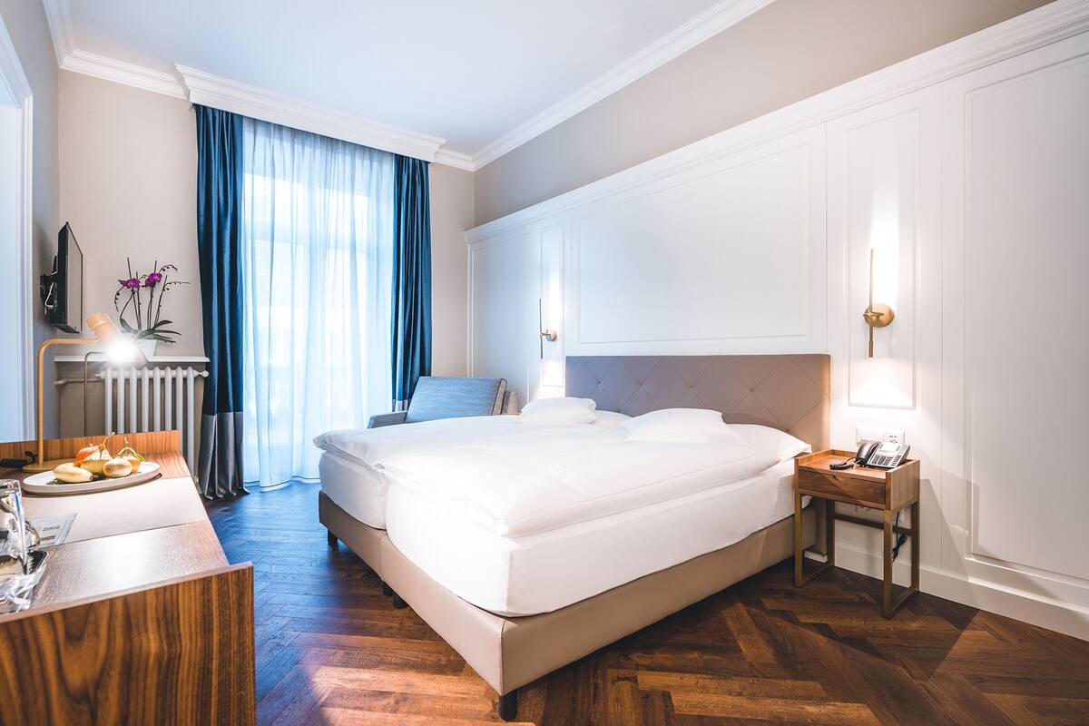 A hotel room with a large bed, a bedside table with a lamp, a desk with a chair, and a view of the outdoors through a window with curtains.