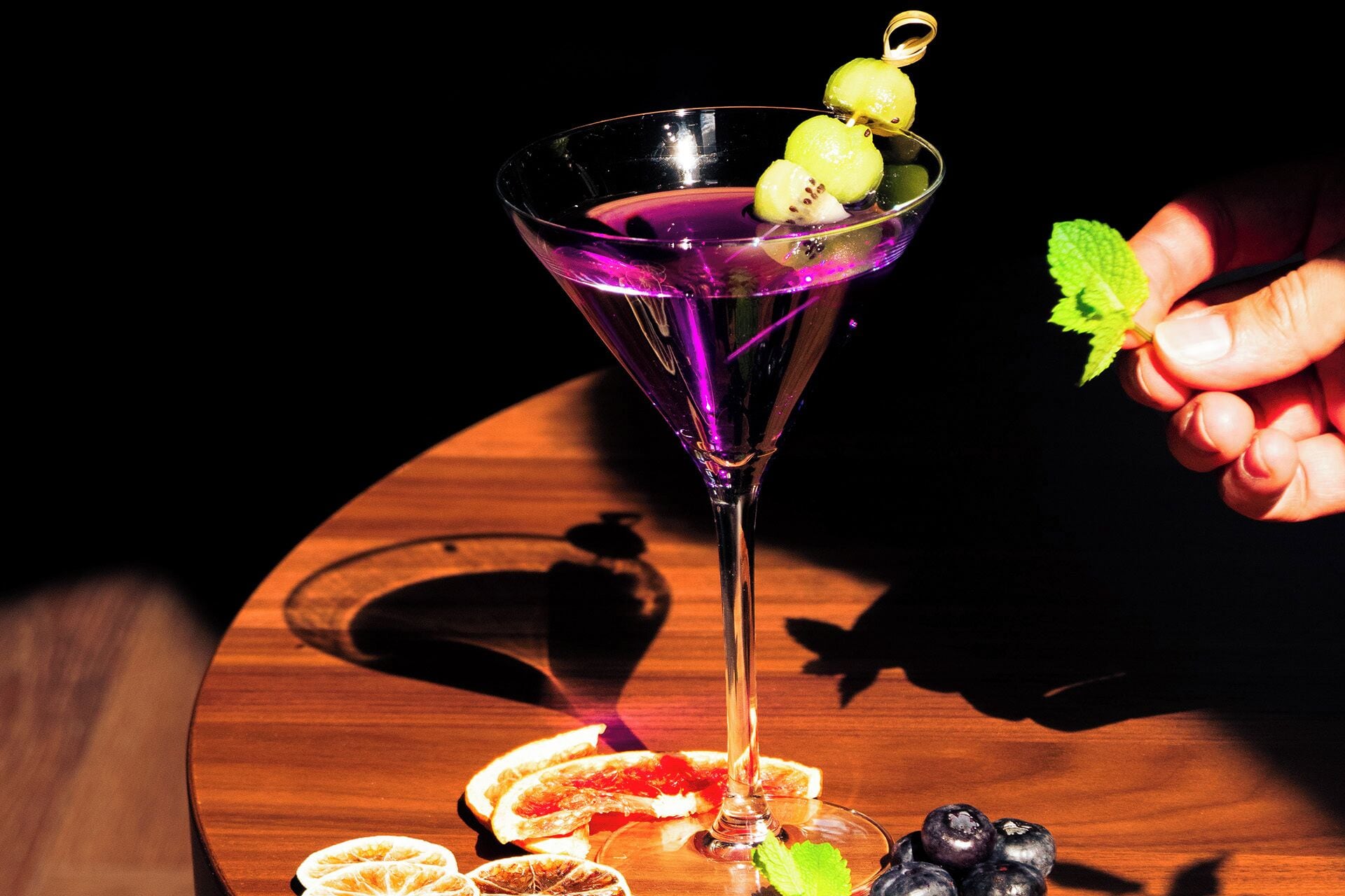 A martini glass with a purple liquid, garnished with a lemon and a mint leaf, on a wooden table with citrus slices and berries.