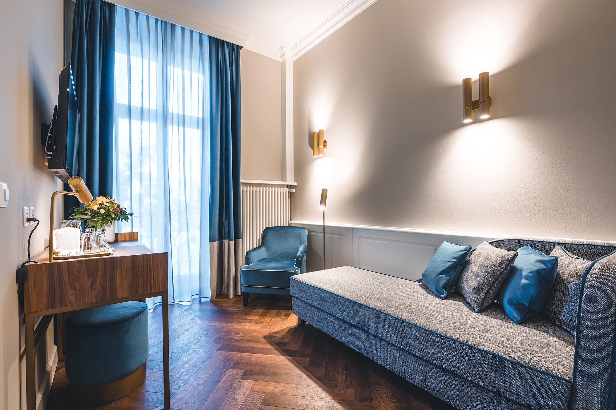 Une chambre d’hôtel moderne avec un plancher en bois, un canapé bleu agrémenté de plusieurs coussins, un bureau avec une lampe et une grande fenêtre ornée de rideaux bleus.