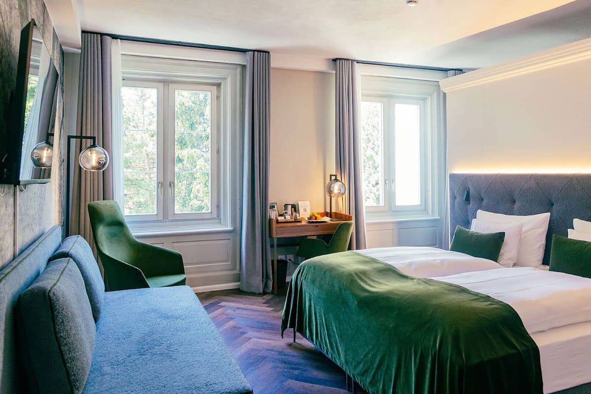 A hotel room with a bed, a desk, a chair, and two windows.