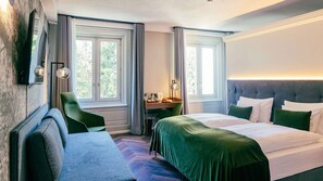 Quarto de hotel com cama, escrivaninha, cadeira e duas janelas.