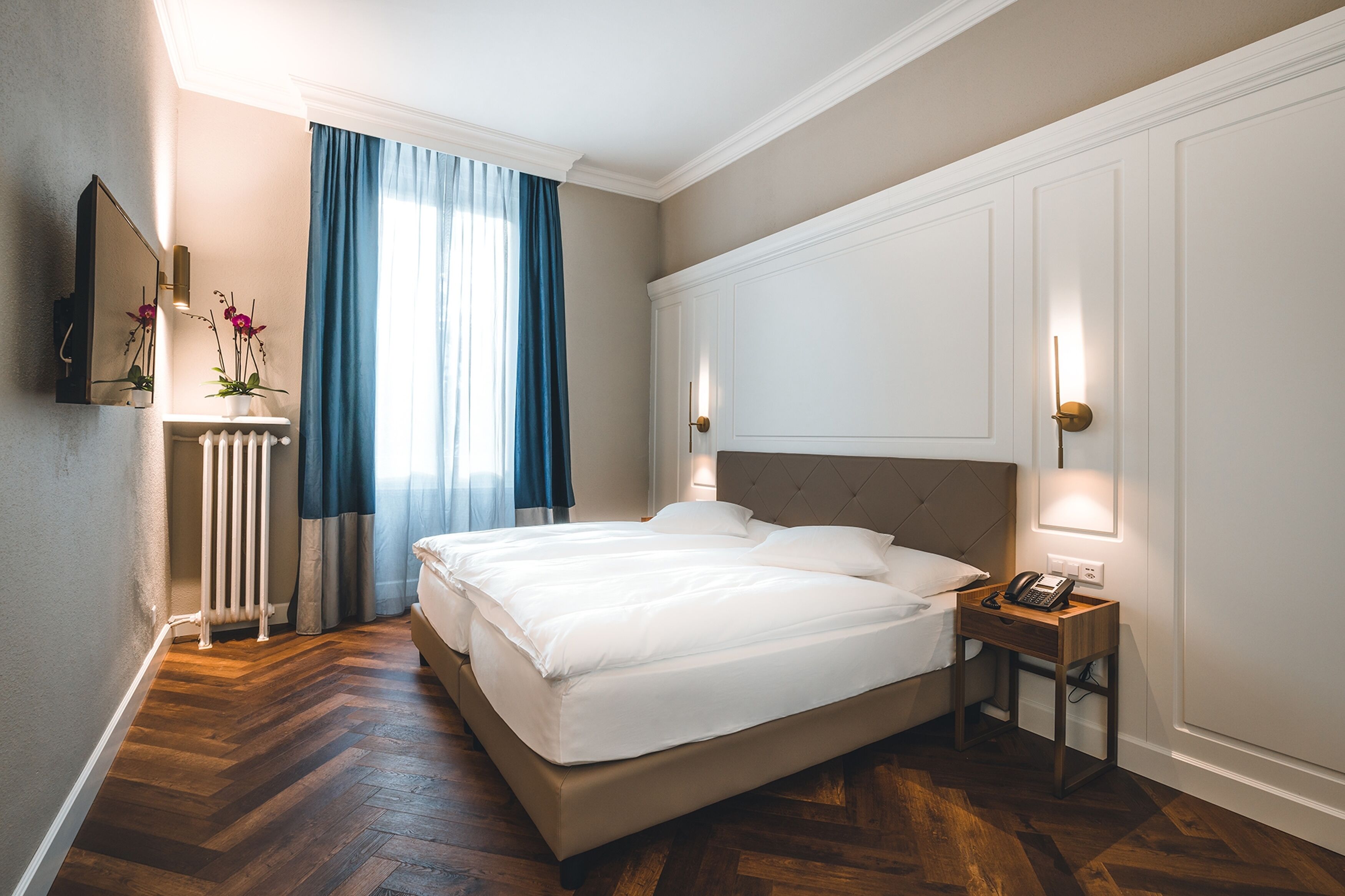 A hotel room with a large bed, a bedside table with a phone, a radiator, and a mirror on the wall.