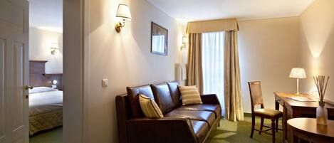 A hotel room with a brown leather sofa, a wooden chair, a small table, and a bed in the background.