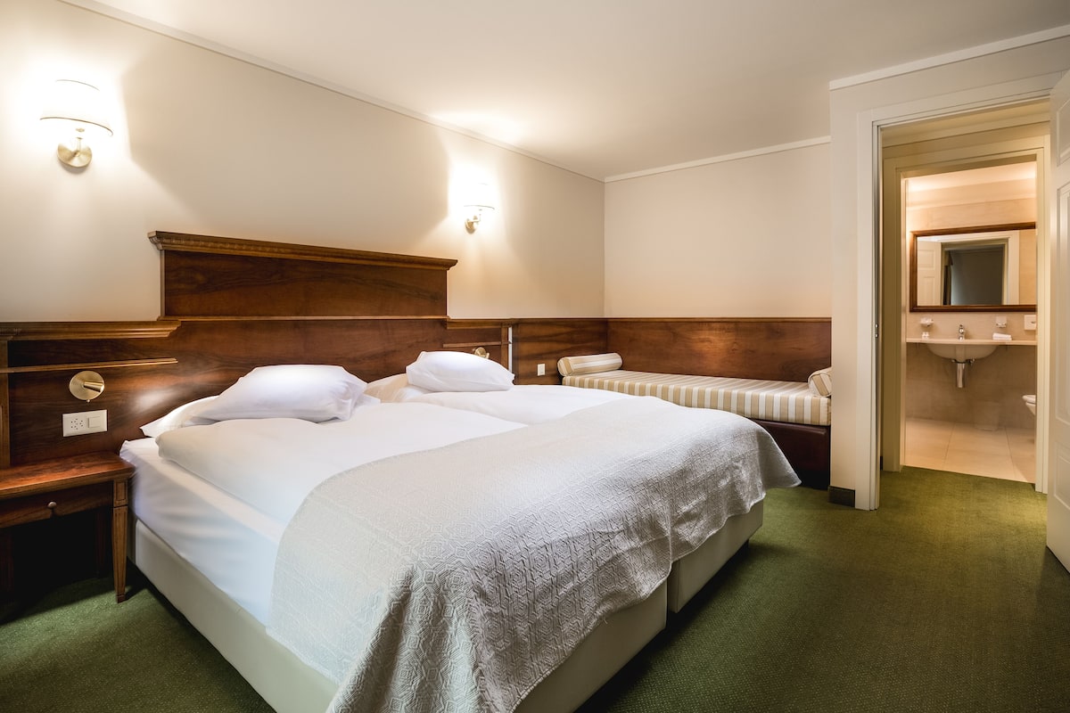 A hotel room with a large bed, two bedside tables, a wooden headboard, a green carpet, and a bathroom visible through an open door.