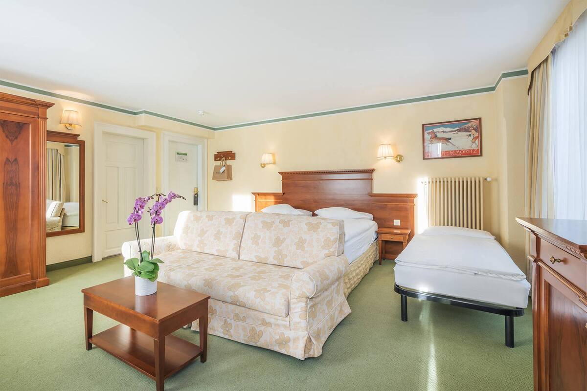 A hotel room with a bed, a sofa, a small table with a vase of flowers, a dresser, and a painting on the wall.