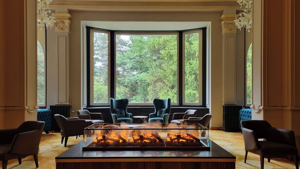 A spacious room with a large fireplace, multiple seating areas, and large windows offering a view of greenery. - Hotel Reine Victoria (St. Moritz)