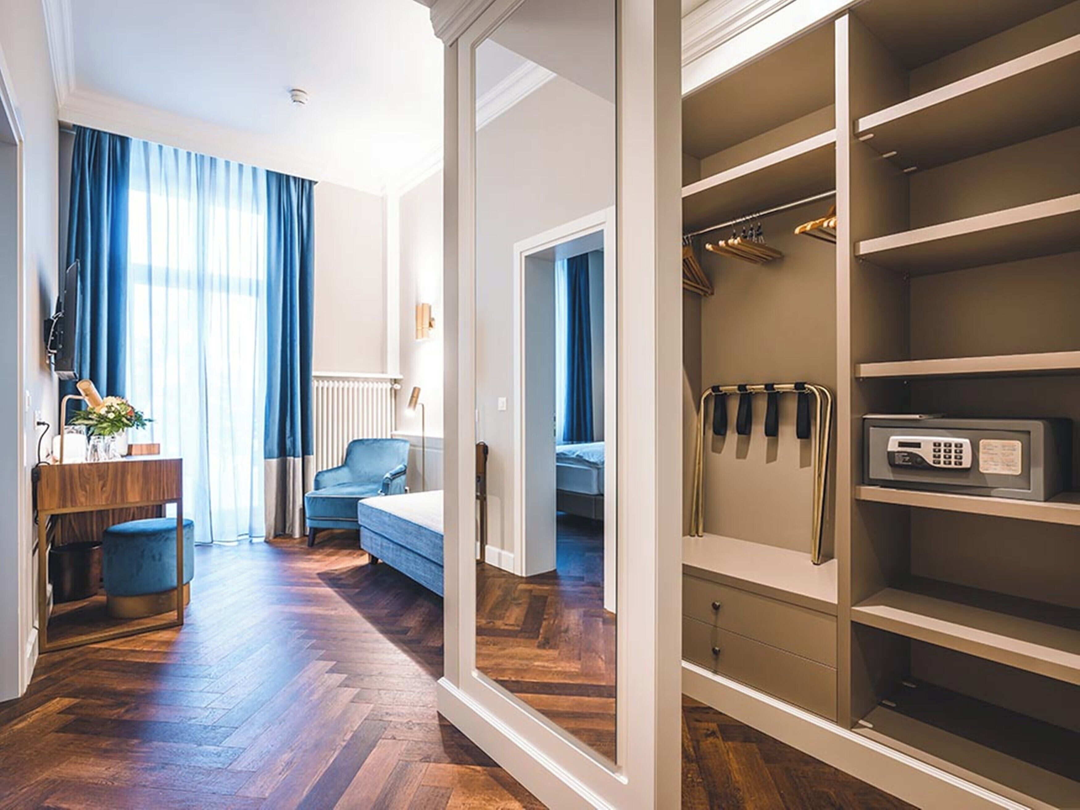 A hotel room with a wooden floor, a desk with a lamp, a chair, a bed, and a walk-in closet with shelves and a safe.