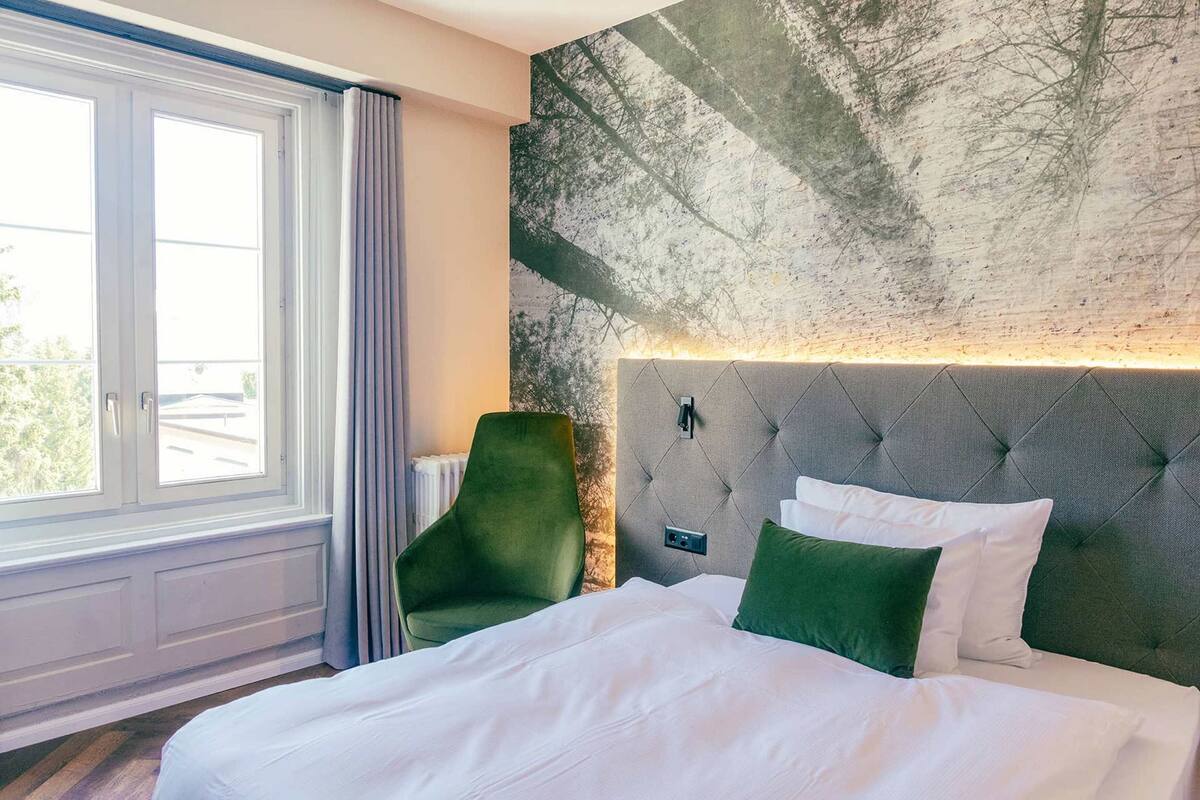 A hotel room with a large bed, a green chair, a window with curtains, and a textured wall.