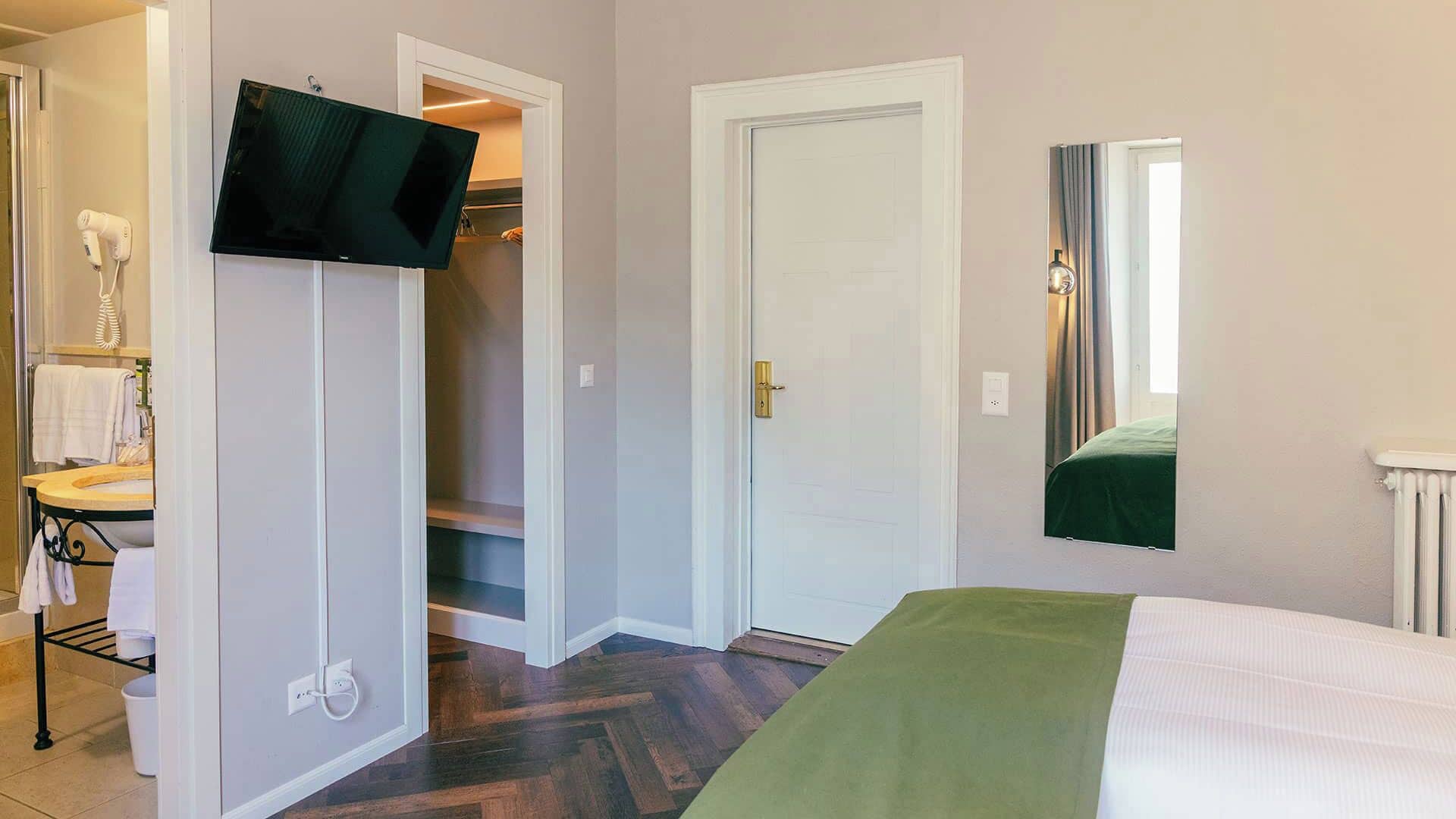 Ein Hotelzimmer mit Bett, einem an der Wand befestigten Fernseher, einem Badezimmer mit Waschbecken und Handtuchhalter sowie einem Schrank mit Tür.