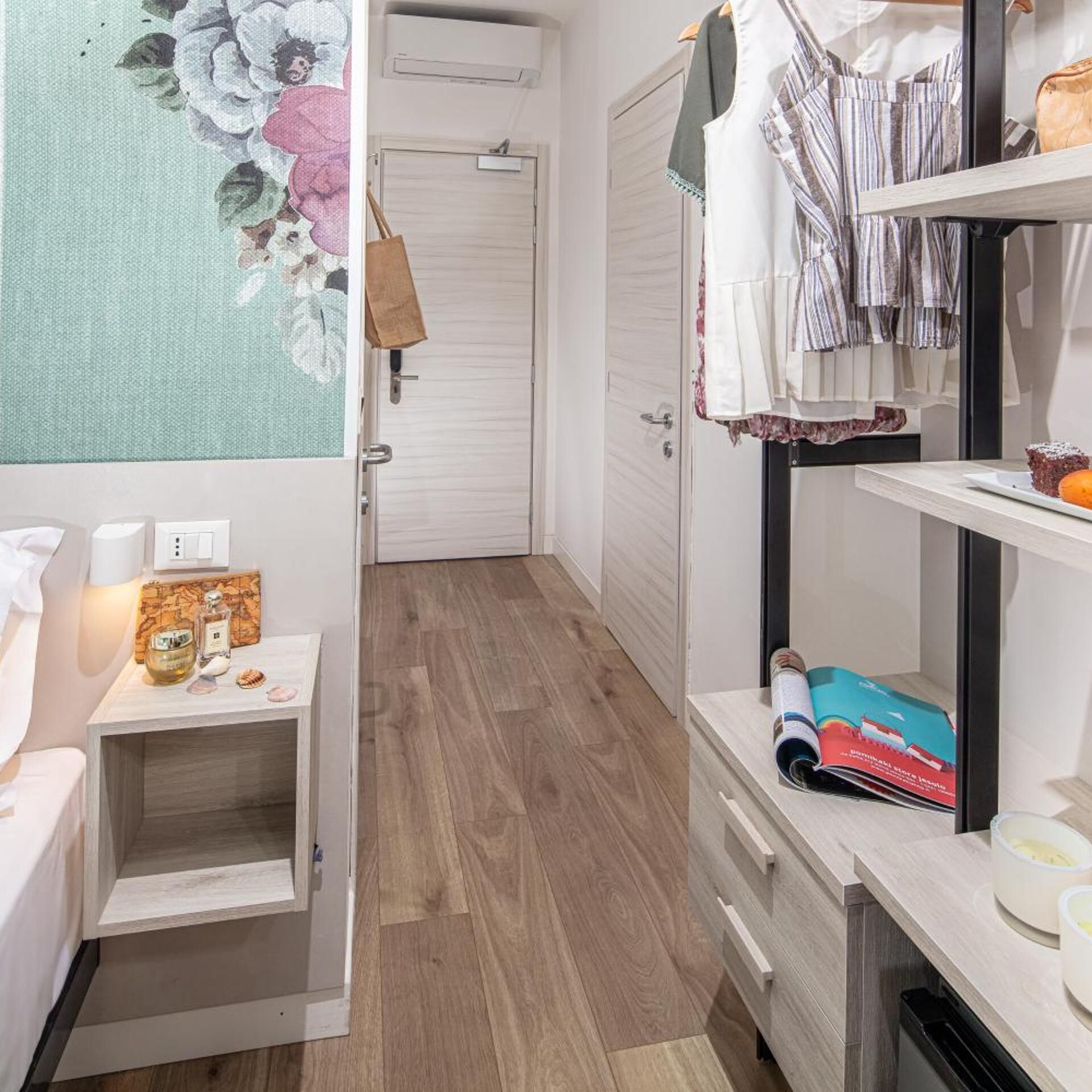 A hotel room with a wooden floor, a bed with white bedding, a vanity with a mirror, a small table with toiletries, and a wardrobe with hanging clothes.
