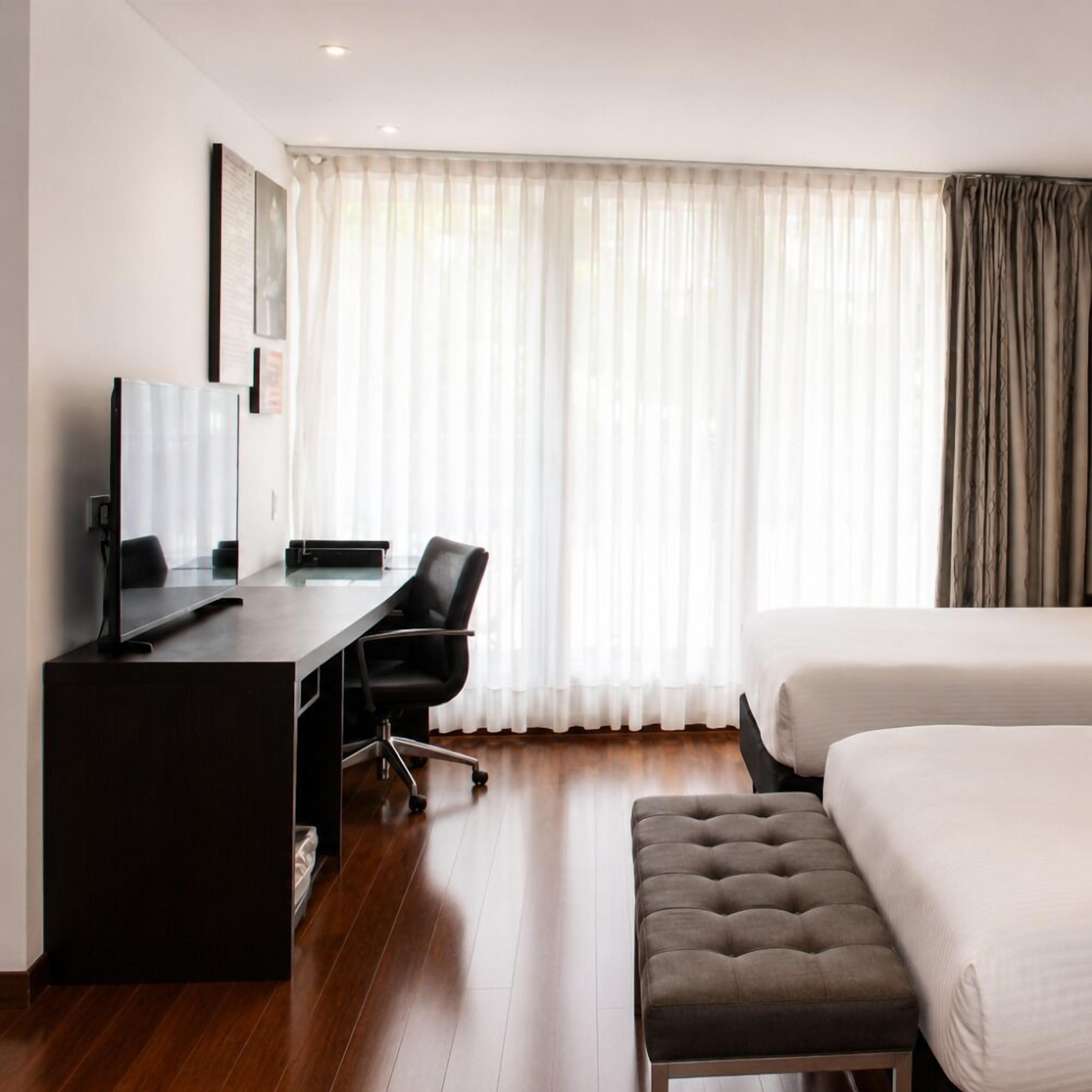 Quarto de hotel com uma cama, uma escrivaninha com computador, uma cadeira e uma janela com cortinas.
