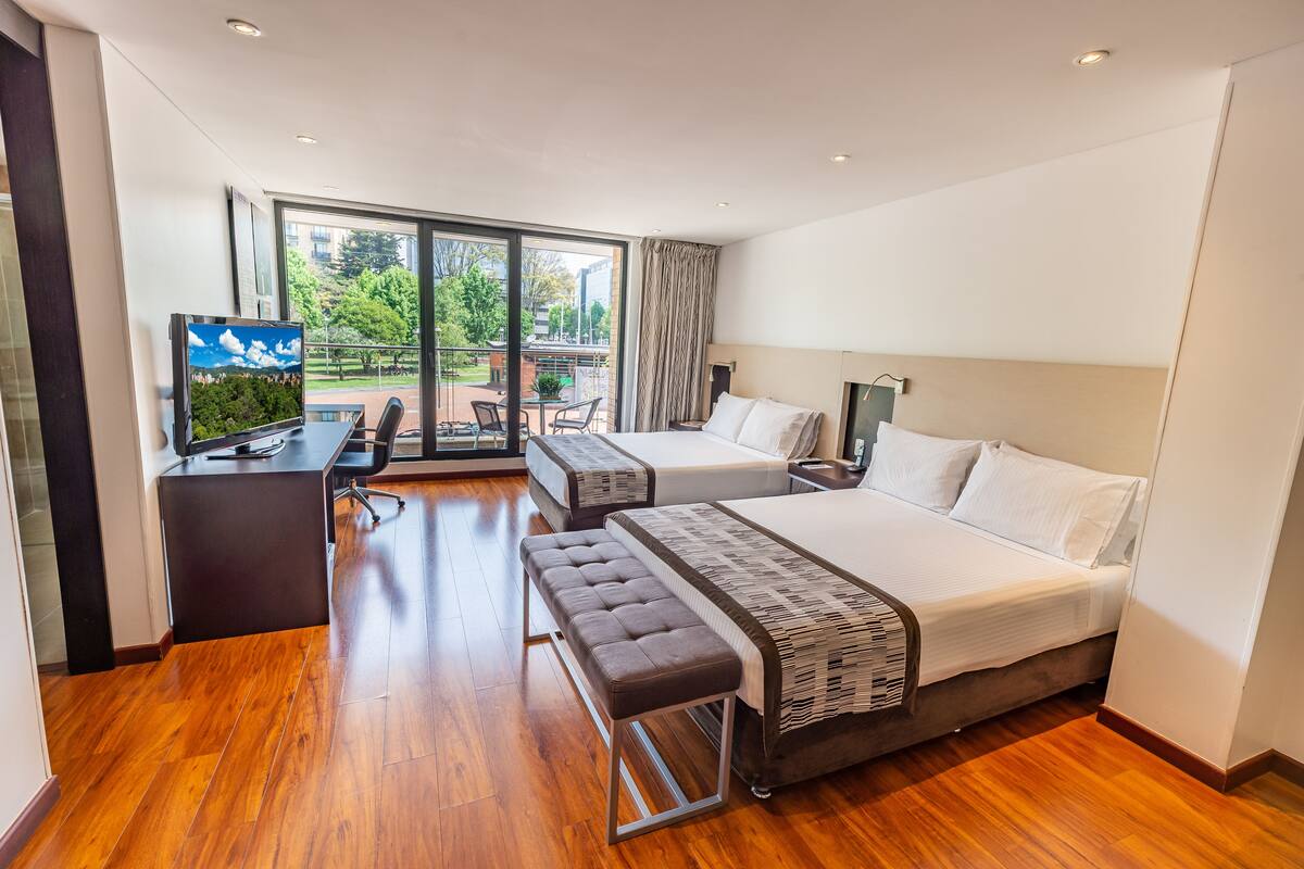 A hotel room with a large bed, a smaller bed, a desk with a TV, and a view of the outdoors.