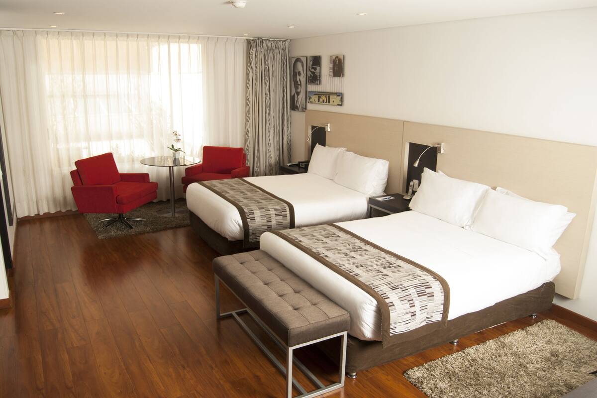 A hotel room with two beds, a red armchair, a small table, and a large window with sheer curtains.