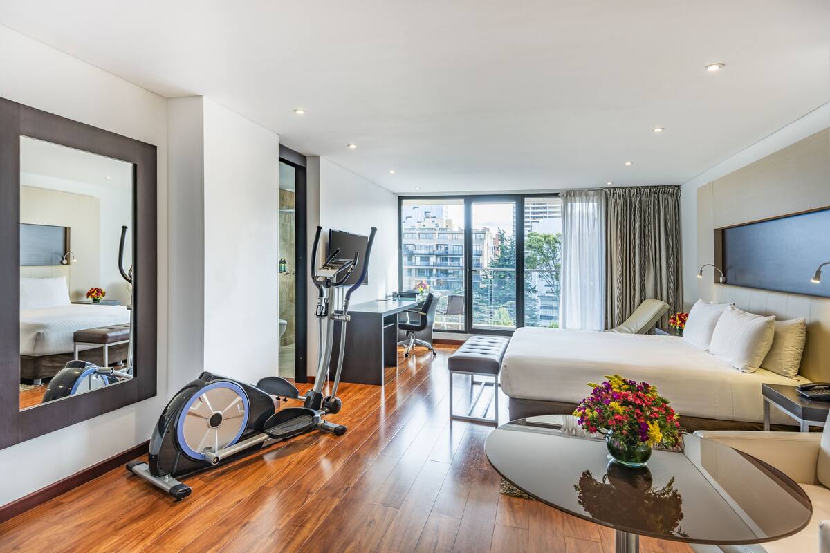 A modern hotel room with a large bed, a desk with a chair, a treadmill, and a large window offering a city view.
