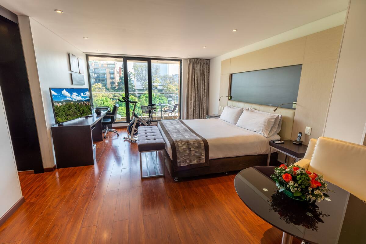 A modern hotel room with a large bed, a desk with a flat-screen TV, a treadmill, and a balcony with a city view.