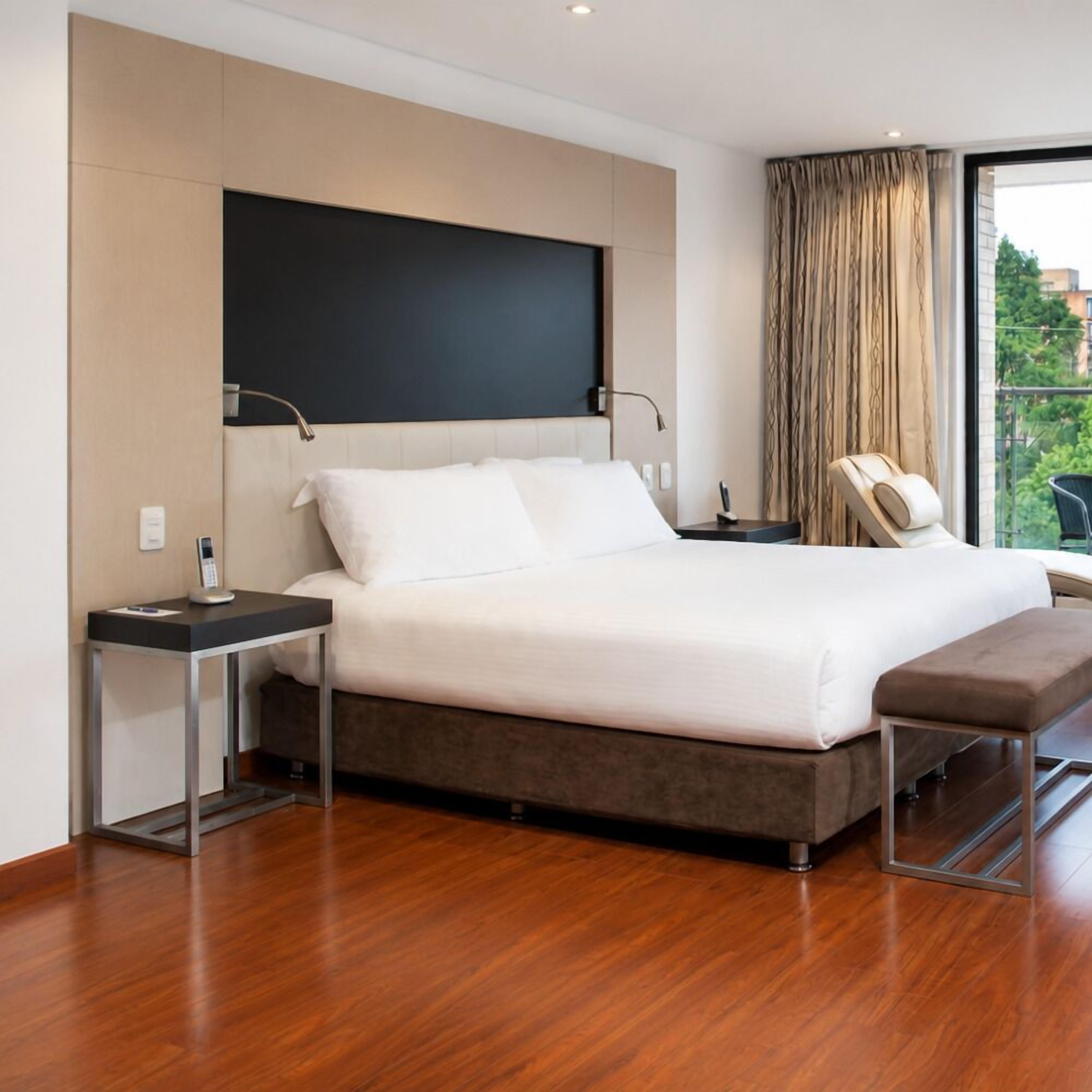 Habitación de hotel moderna con una cama grande, mesitas de noche, un televisor de pantalla plana, un sillón de masajes y vistas a zonas verdes.