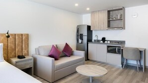 A compact hotel room with a bed, a sofa, a small dining table, a kitchenette, and a wooden headboard.