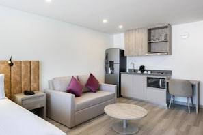 Una habitación de hotel compacta con cama, sofá, pequeña mesa de comedor, kitchenette y cabecero de madera.