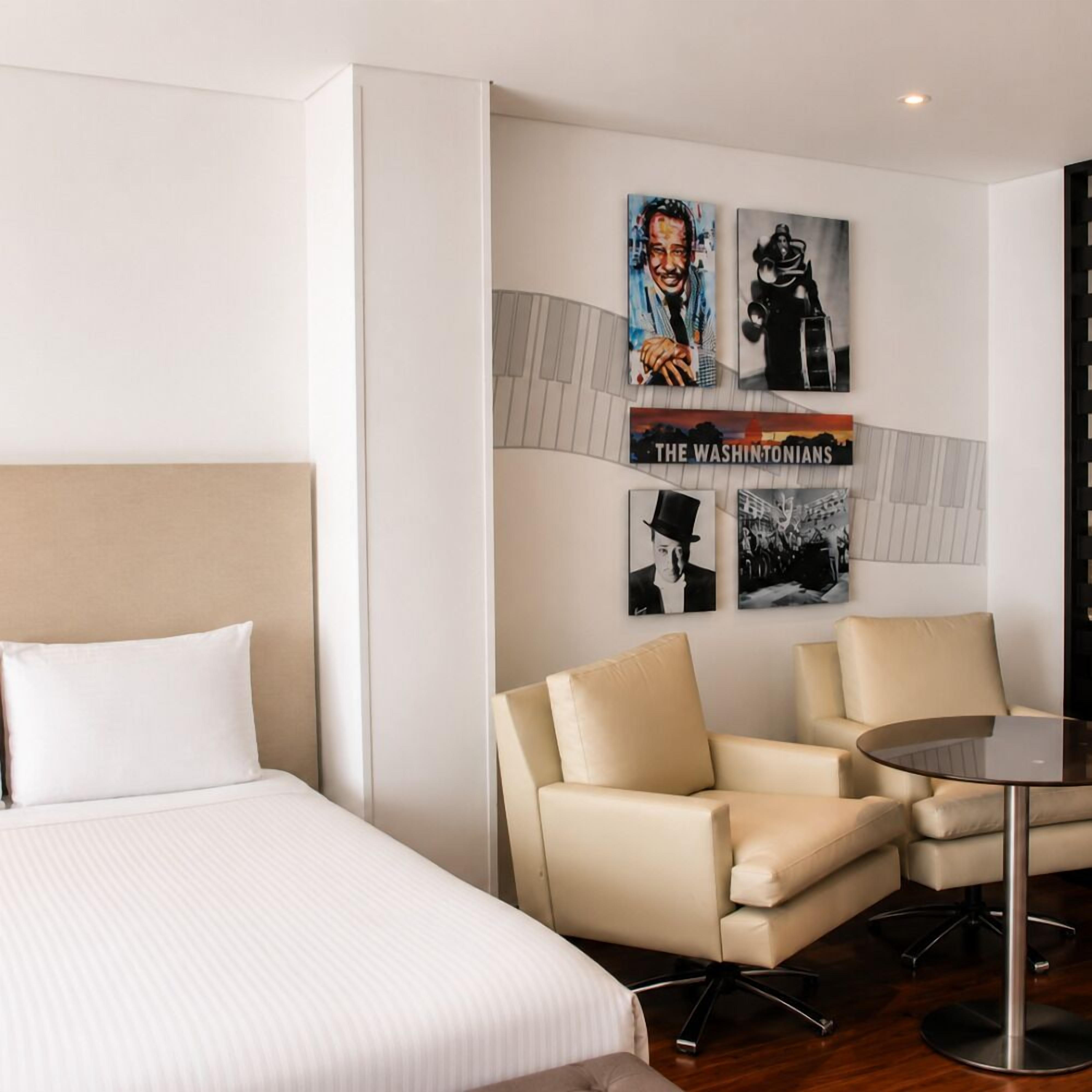 A hotel room with a bed, two armchairs, a small table, and wall art including a poster titled 'THE WASHINGTONIANS'.