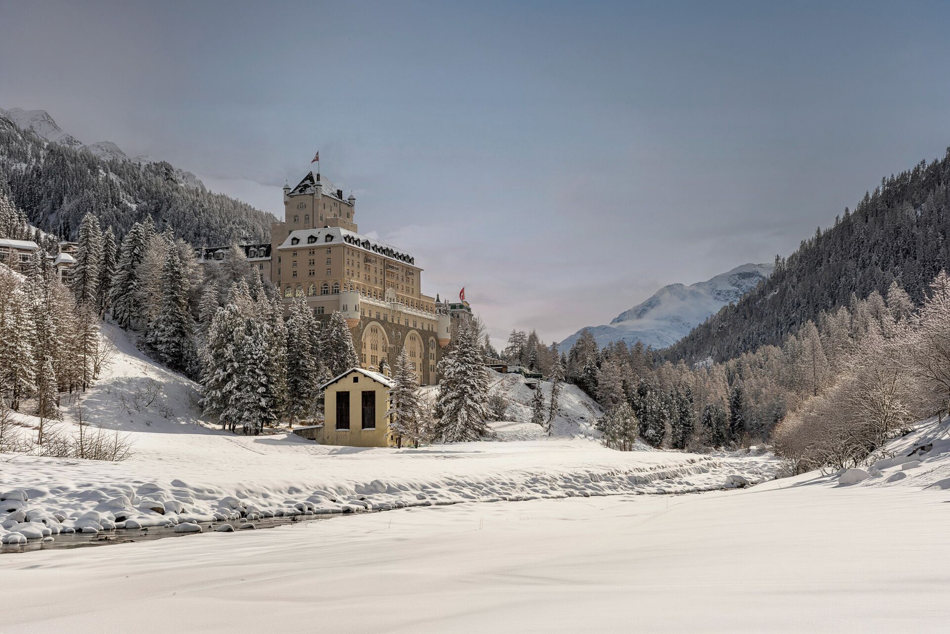 A historic building with a prominent tower, surrounded by snow-covered trees and mountains.