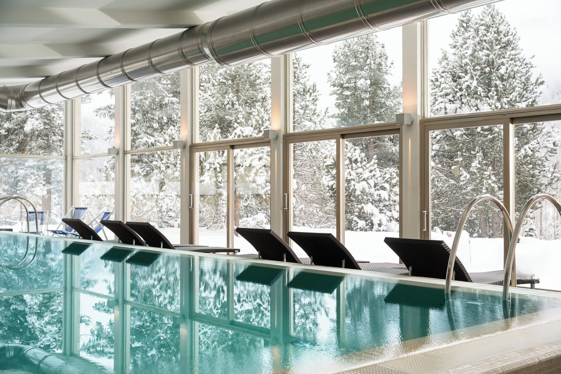 An indoor swimming pool with clear glass walls, multiple lounge chairs, and a view of snow-covered trees outside.