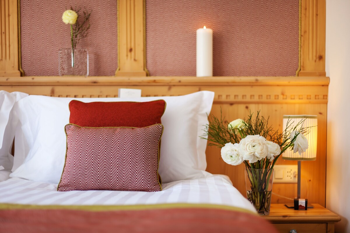 A neatly made bed with white and red pillows, a vase with white flowers, and a lit candle on a wooden nightstand.
