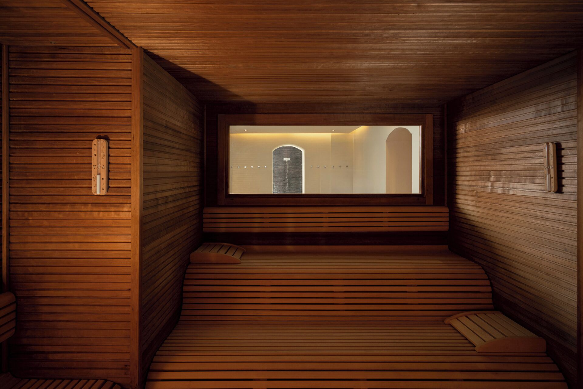 A sauna room with wooden walls and benches, a window with a view of a building, and a thermometer on the wall.