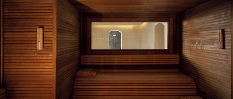 A sauna room with wooden walls and benches, a window with a view of a building, and a thermometer on the wall.