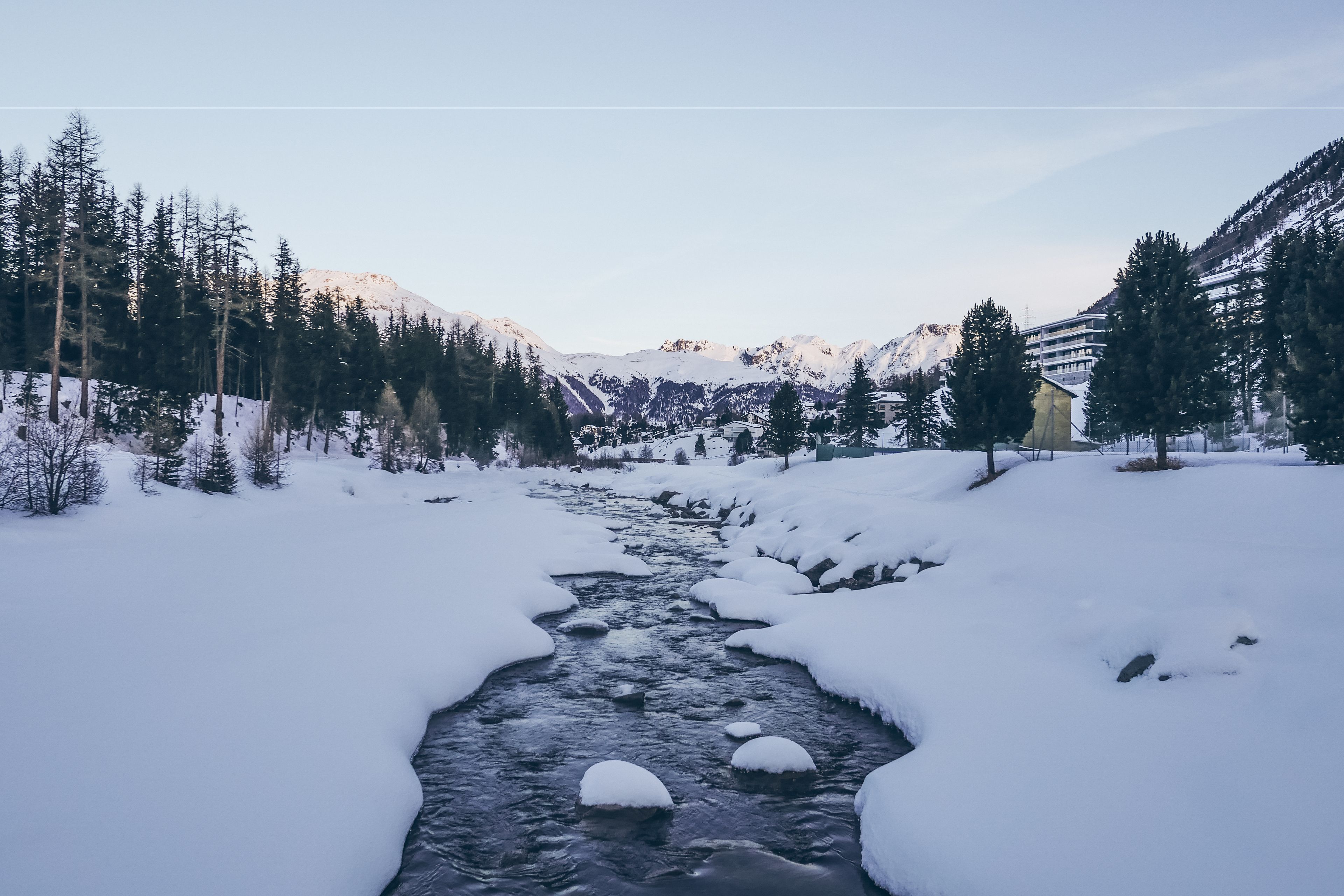 A snow-covered river with mountains in the background and a building on the right.