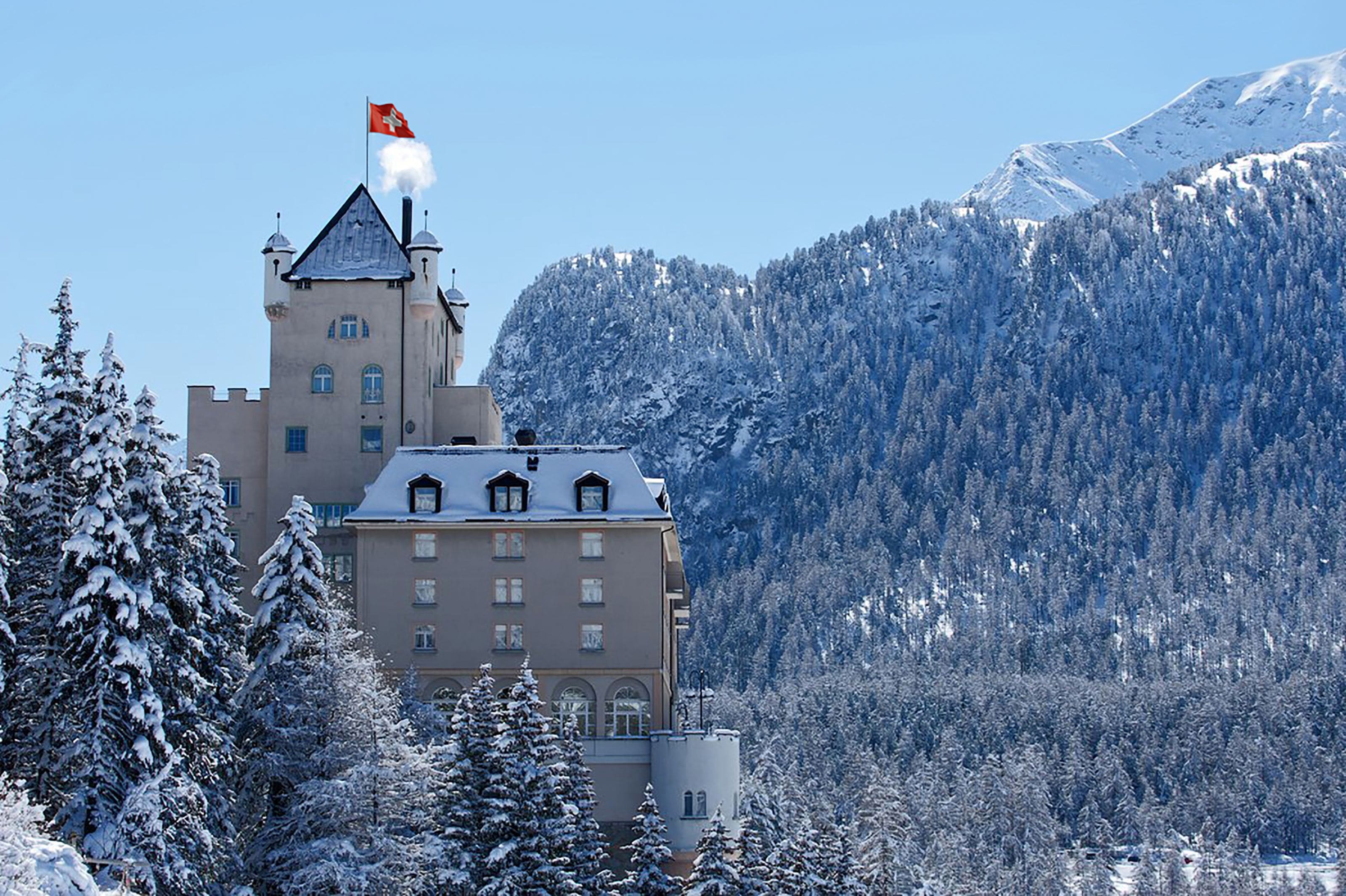 A castle-like building with a tower and flag, surrounded by snow-covered trees and mountains.