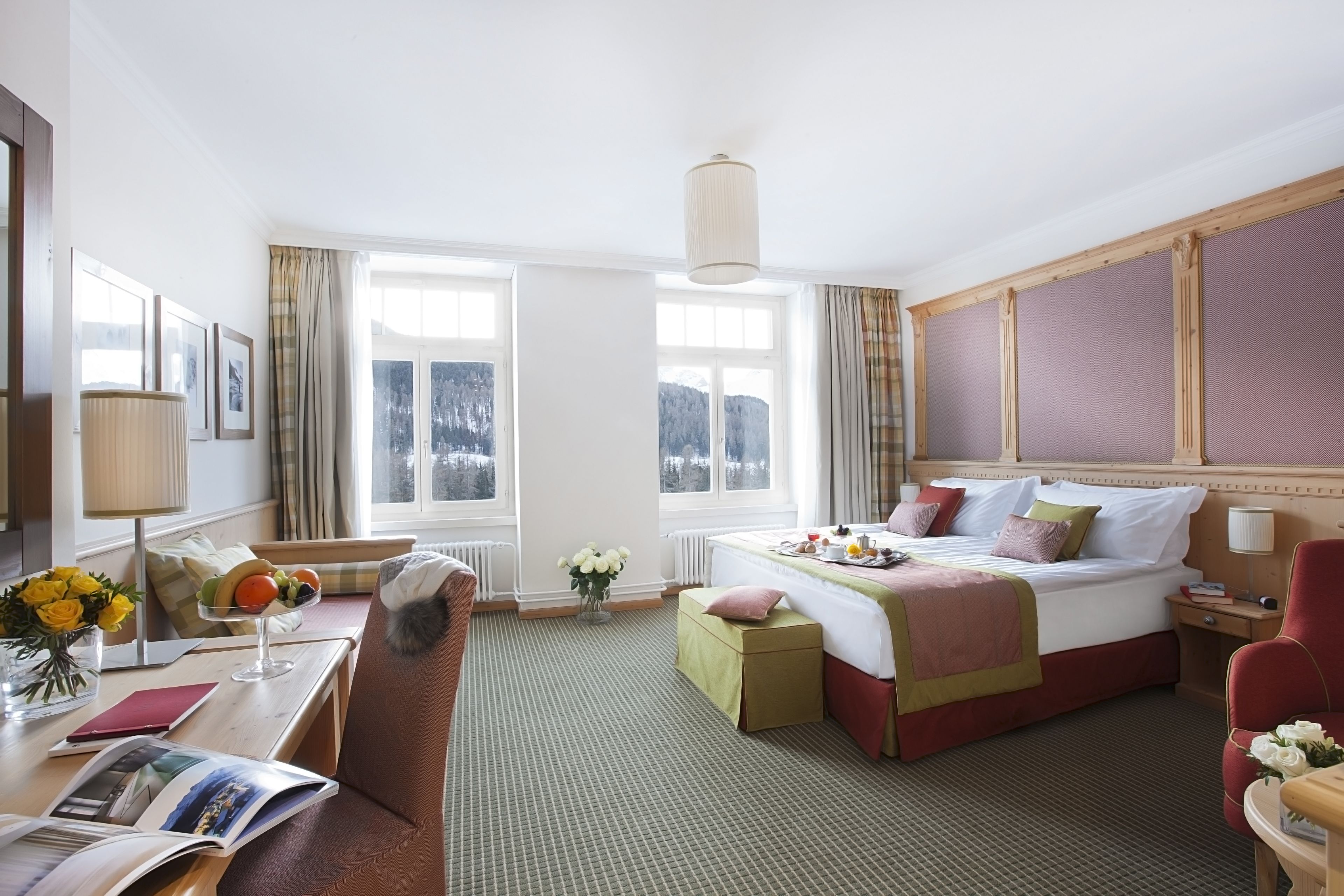 A hotel room with a large bed, a desk with a lamp, a chair, a window with curtains, and a vase with flowers.