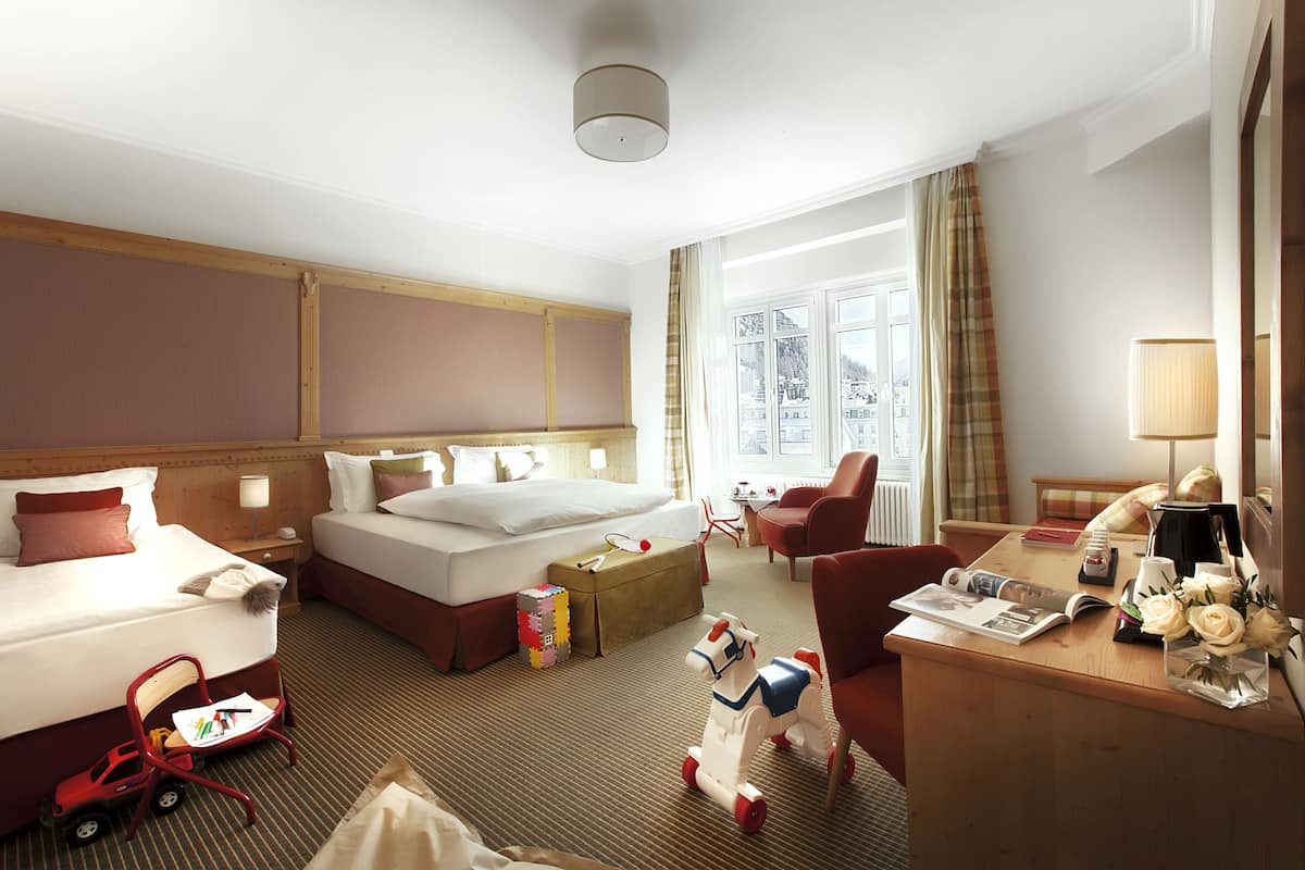 Une chambre d’hôtel avec deux lits, un bureau avec une lampe, une chaise, une petite table avec un vase de fleurs et une fenêtre donnant sur des immeubles.