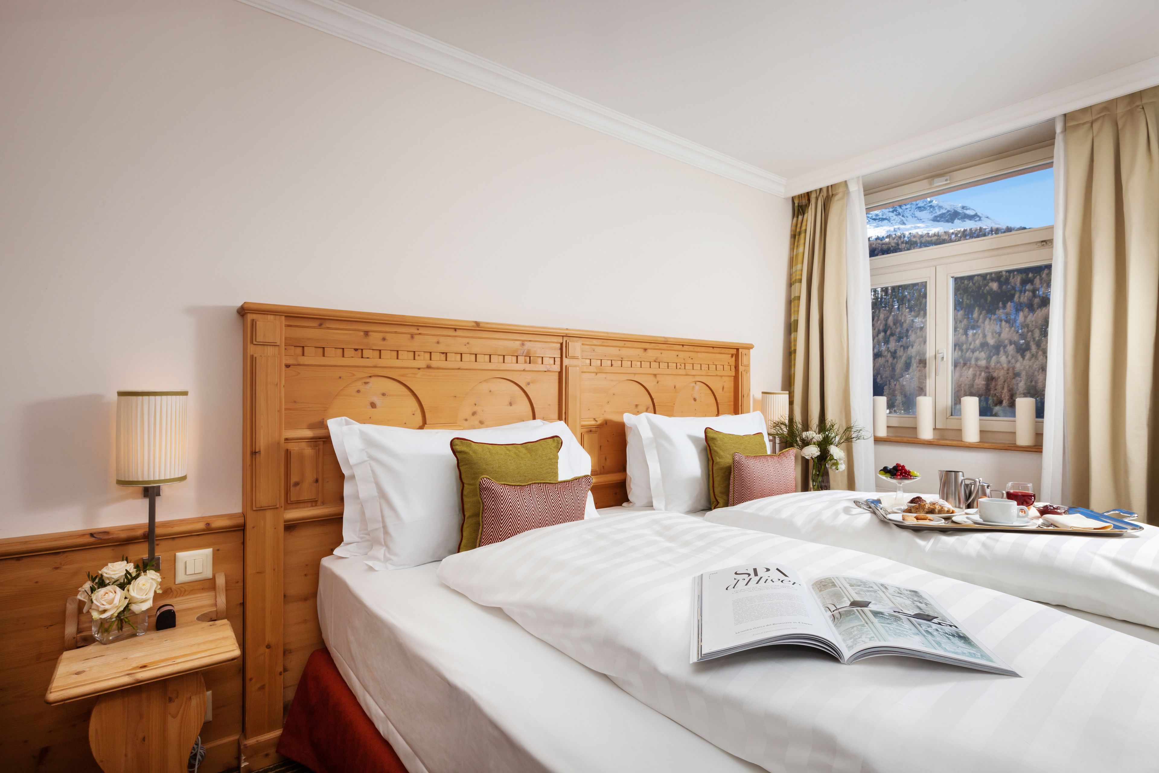 Ein Hotelzimmer mit zwei Betten, einem hölzernen Kopfteil, einem Nachttisch mit Lampe und einem Fenster mit Blick auf einen verschneiten Berg.