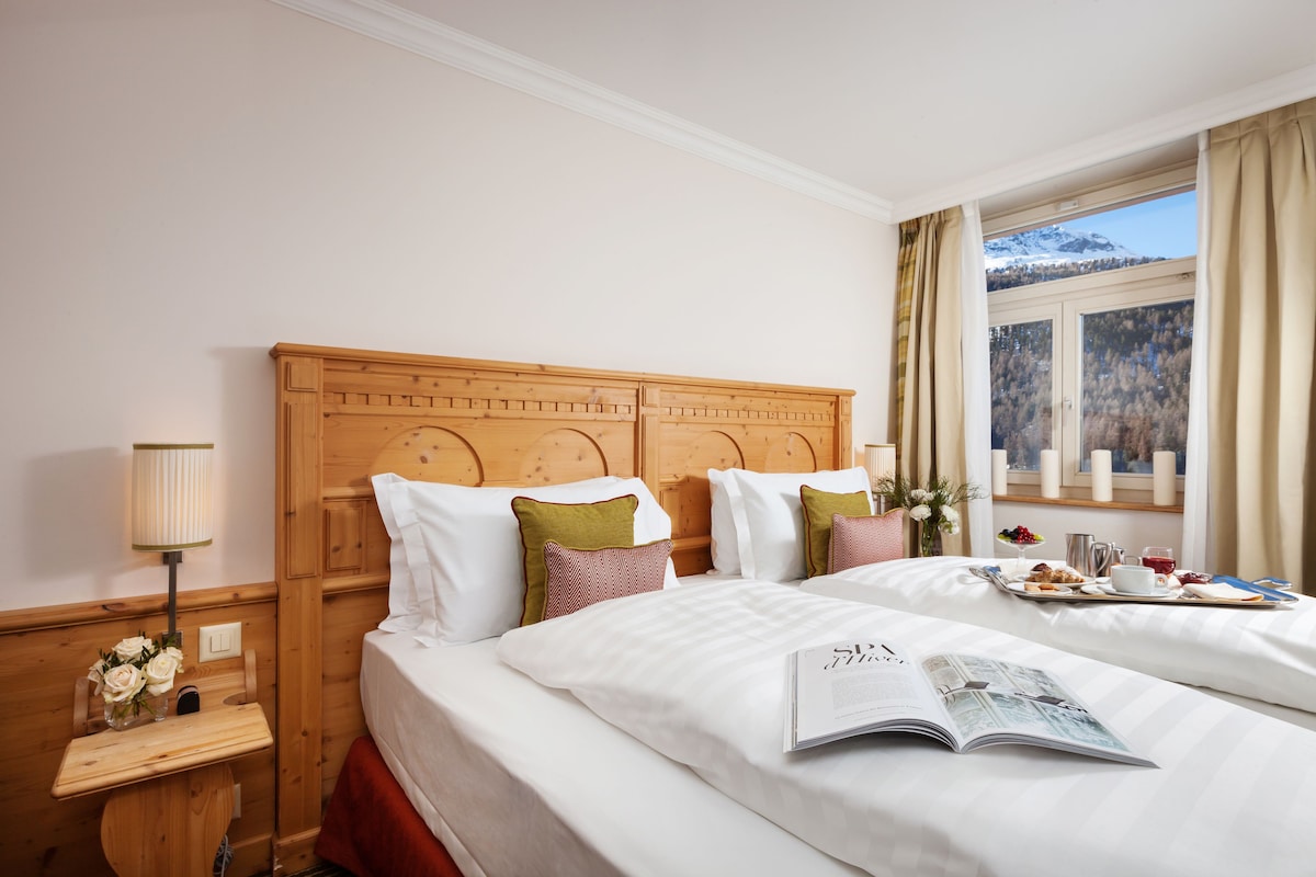 A hotel room with two beds, a wooden headboard, a bedside table with a lamp, and a window with a view of a snowy mountain.