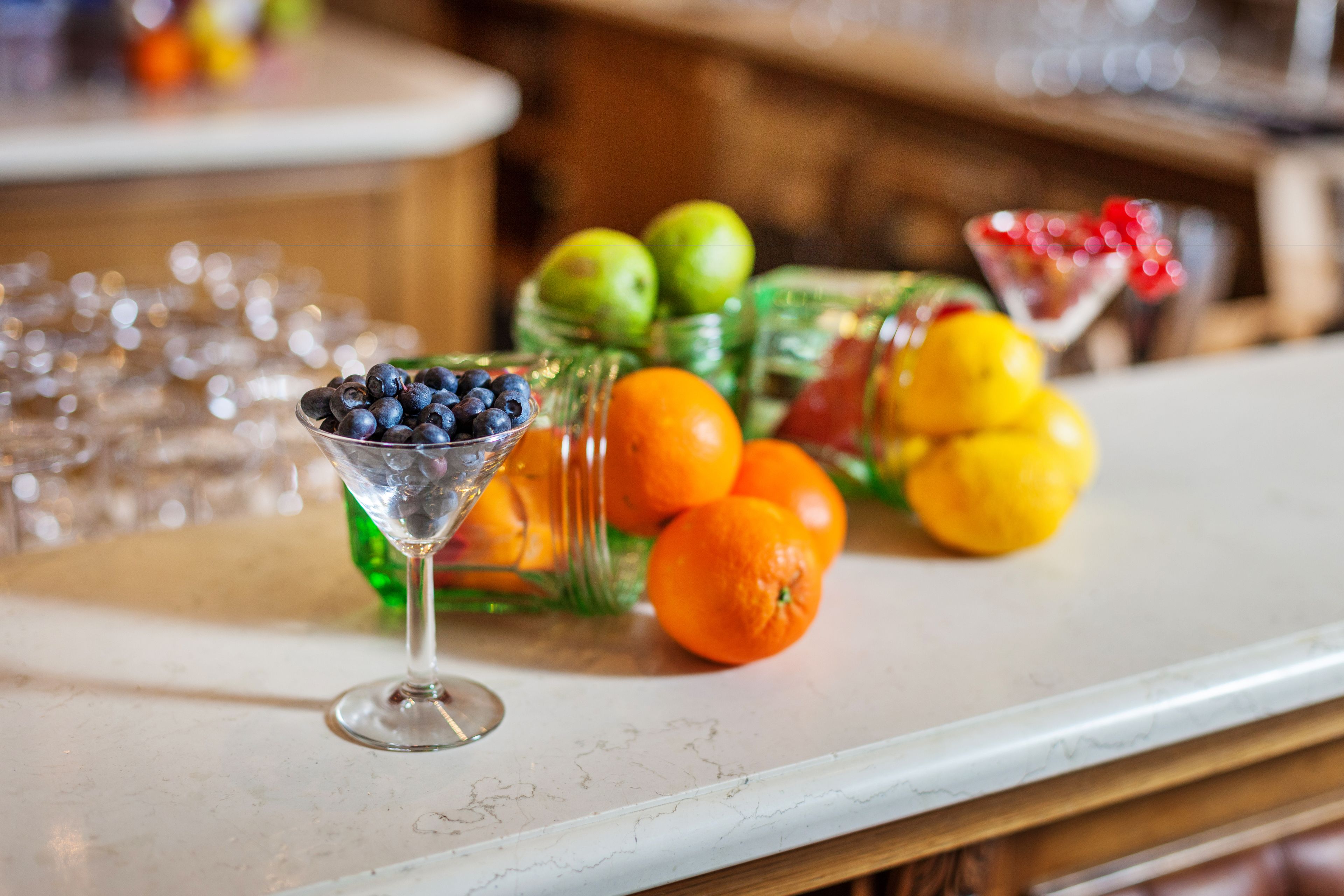 A bar area with a martini glass filled with blueberries, a glass of orange juice, and a bowl of oranges.