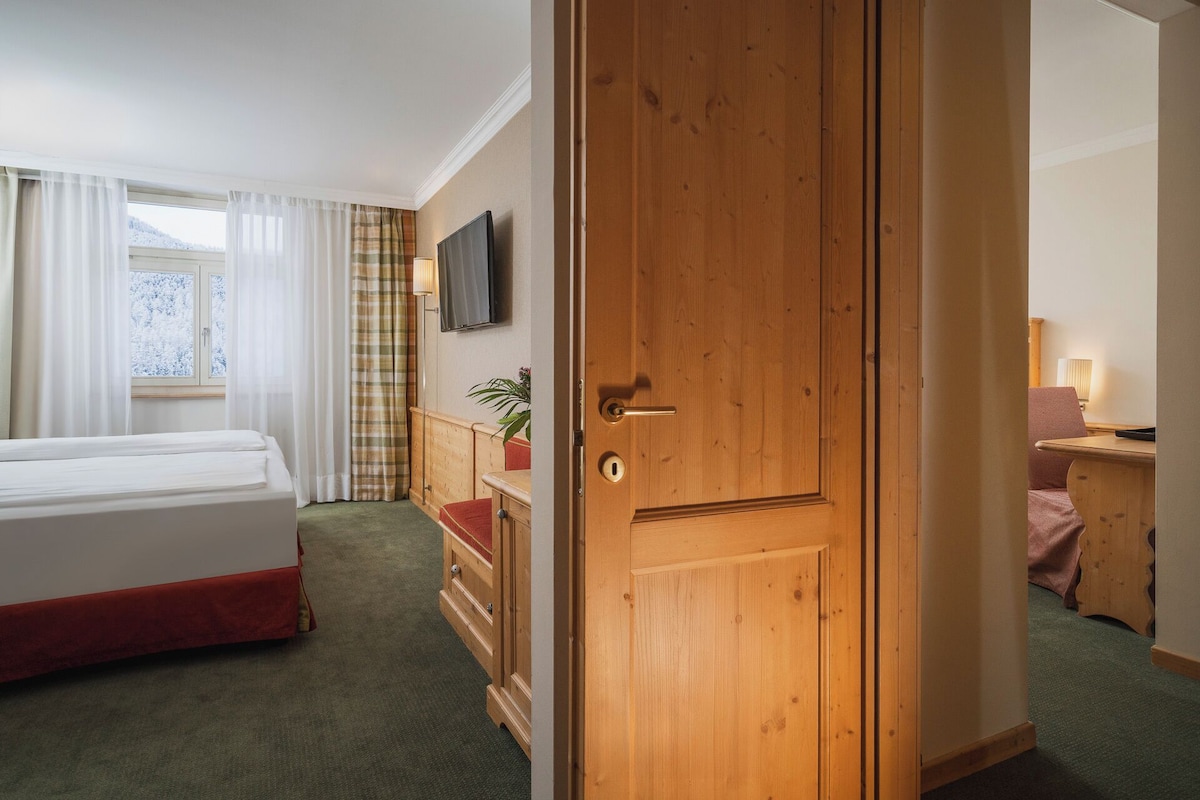 A hotel room with a bed, a television, a wooden door, a desk with a lamp, and a view of snow-covered trees through the window.