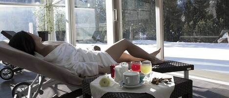 A person lying on a reclining chair by a window with a table in front of them, containing a cup, a glass of juice, and a small plant.