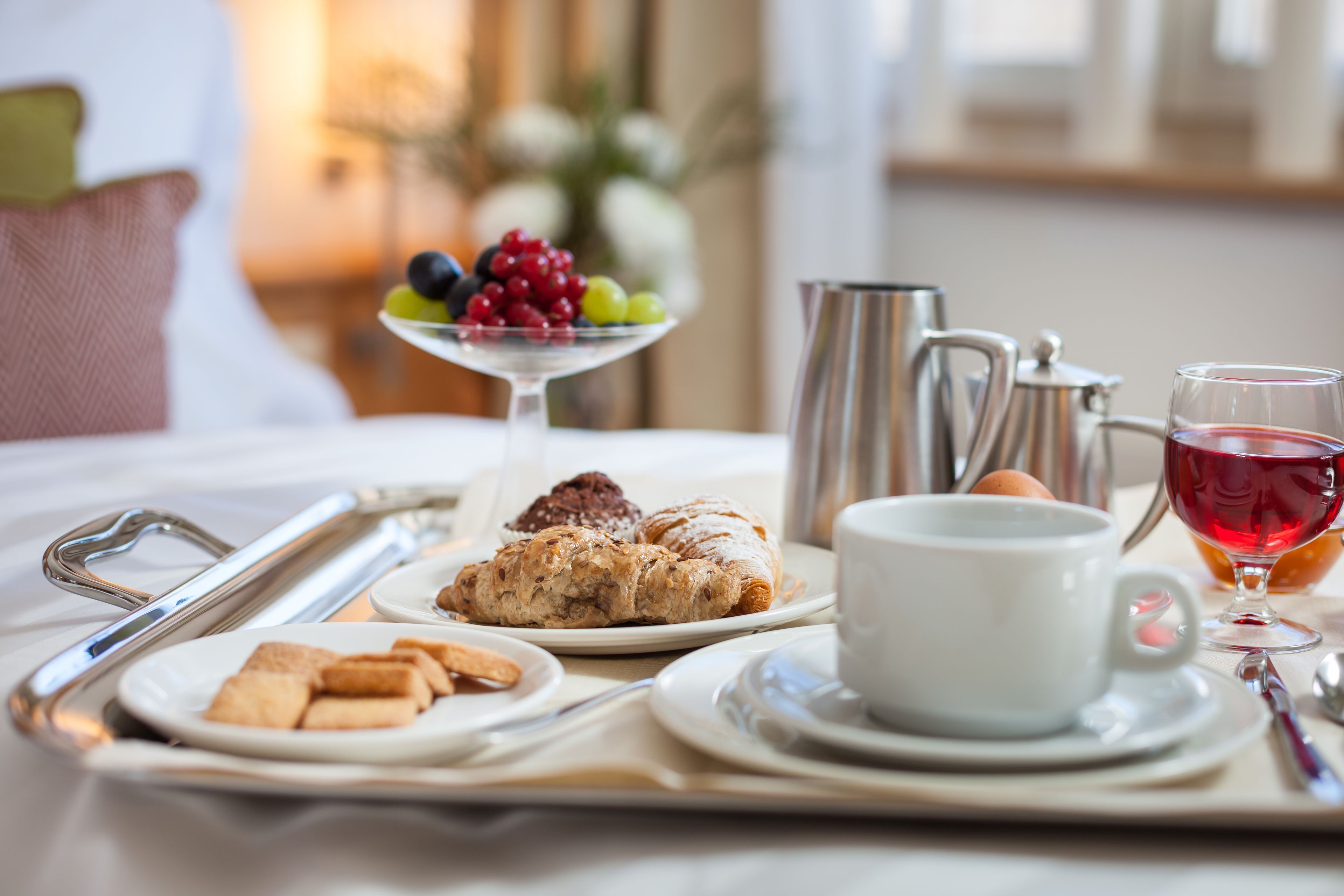 Ein Frühstücksbrett mit Gebäck, Früchten, einer Kaffeekanne, einer Tasse und einem Glas roter Flüssigkeit auf einem Bett.