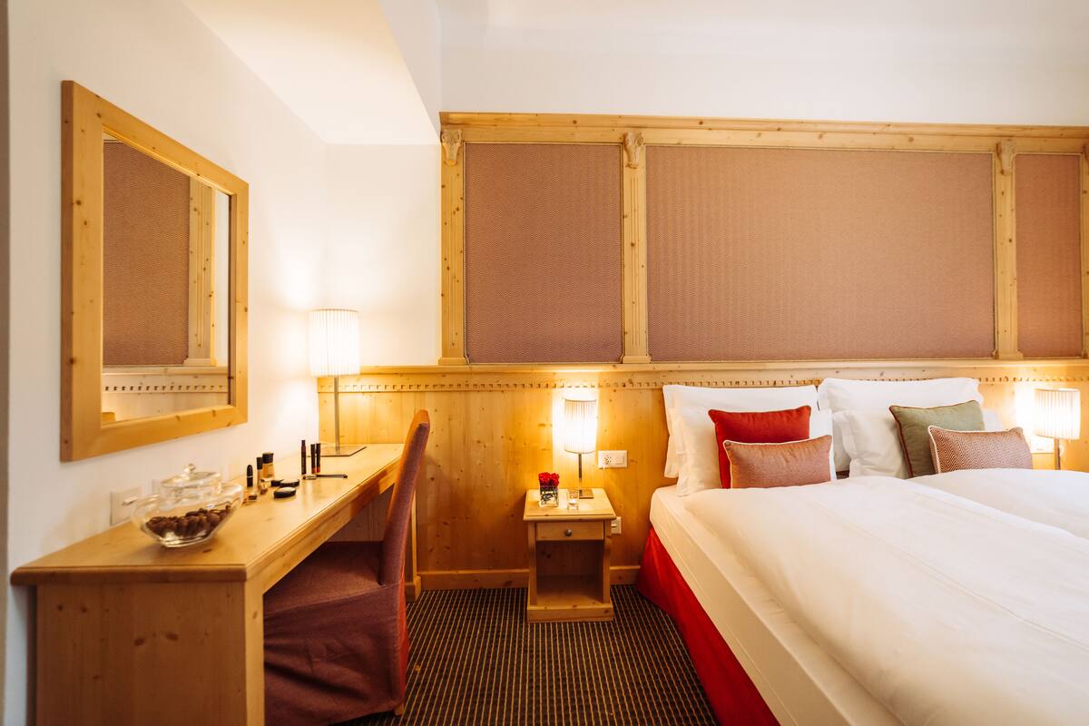 Une chambre d’hôtel avec un bureau en bois, un lit avec plusieurs oreillers, une table de chevet avec une lampe et un miroir au mur.