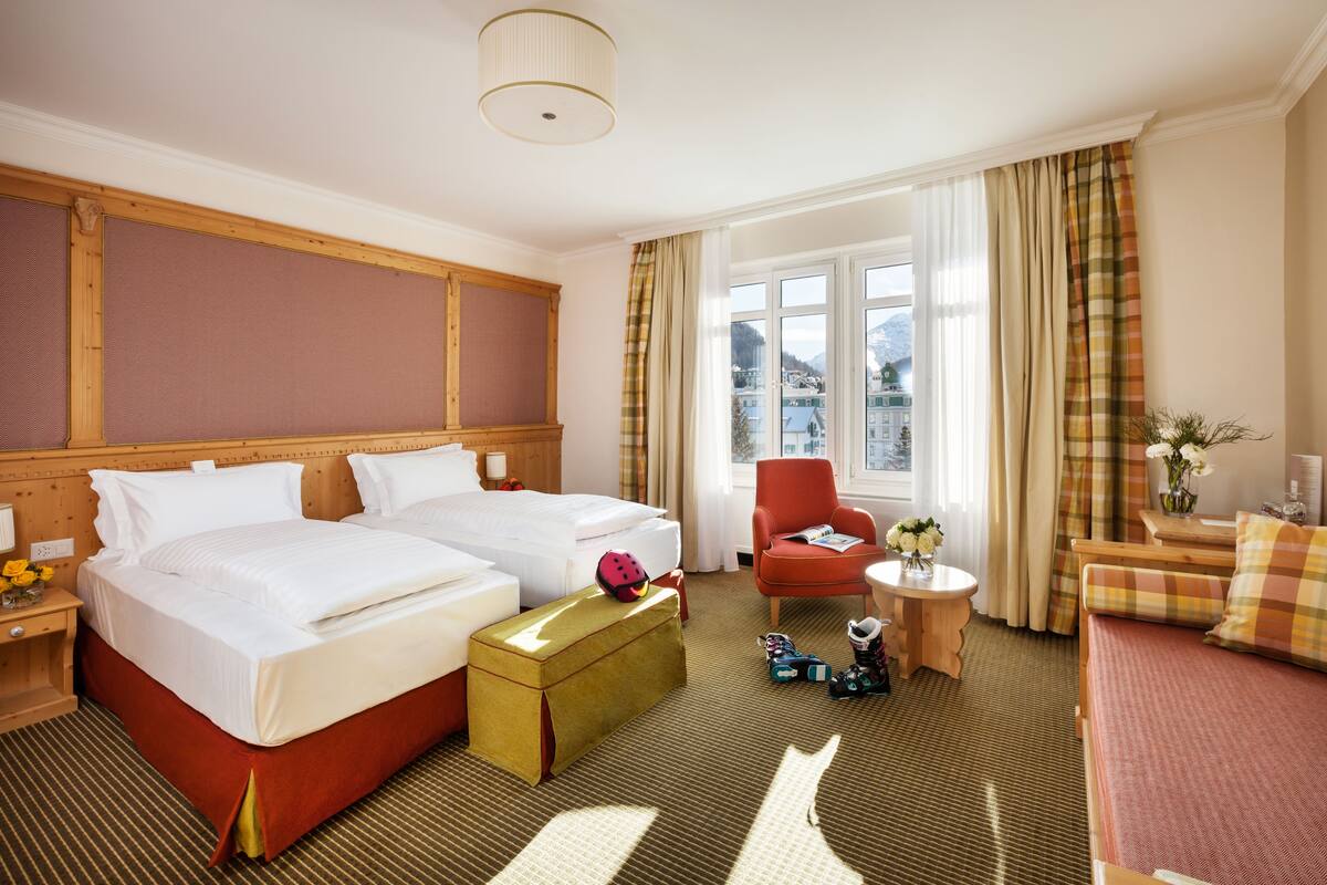 Une chambre d’hôtel avec deux lits, un fauteuil rouge, une petite table sur laquelle est posé un vase de fleurs, et une vue sur une montagne par la fenêtre.