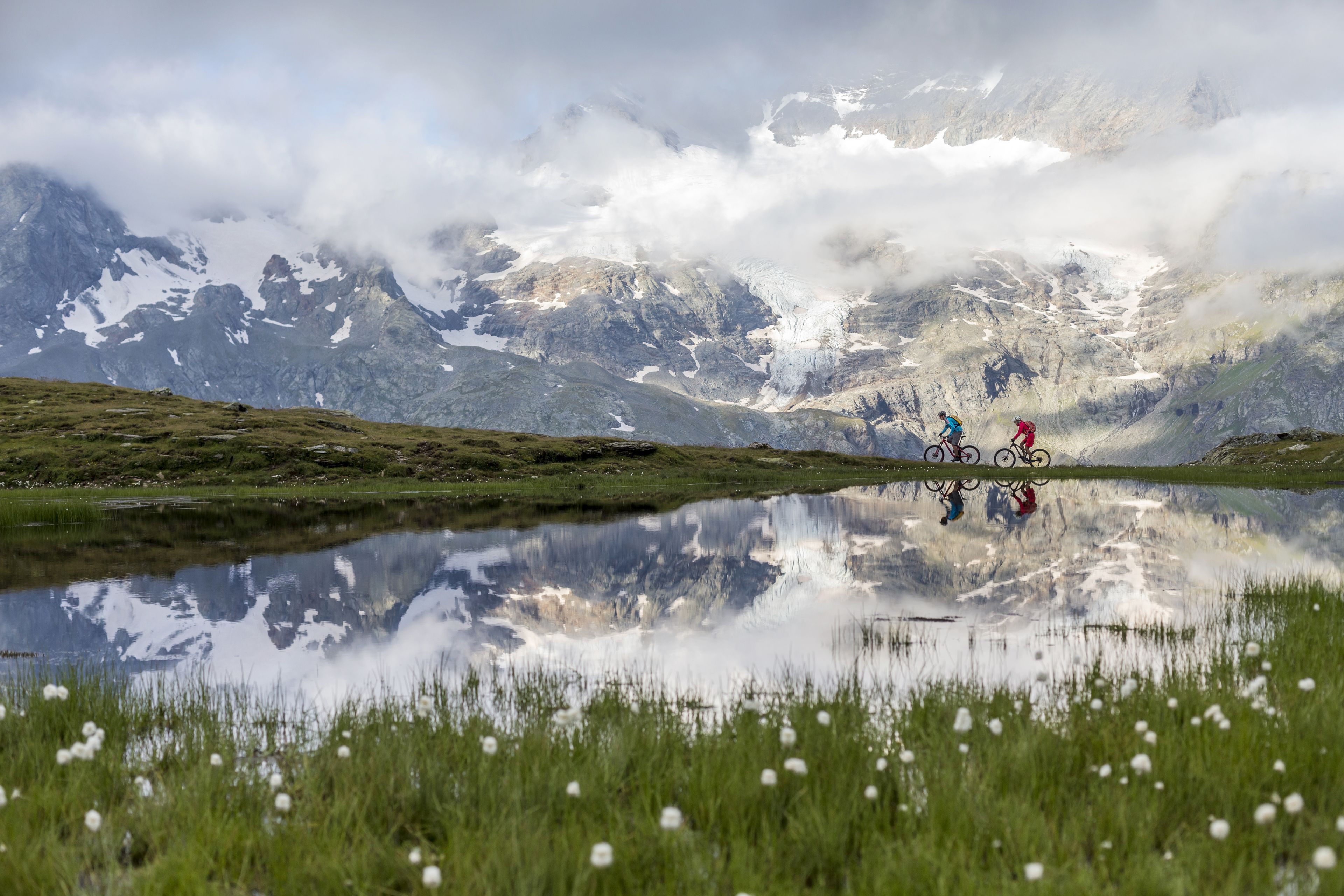 Paesaggio montano con un lago che riflette l'ambiente circostante, inclusi ciclisti e una persona in lontananza.