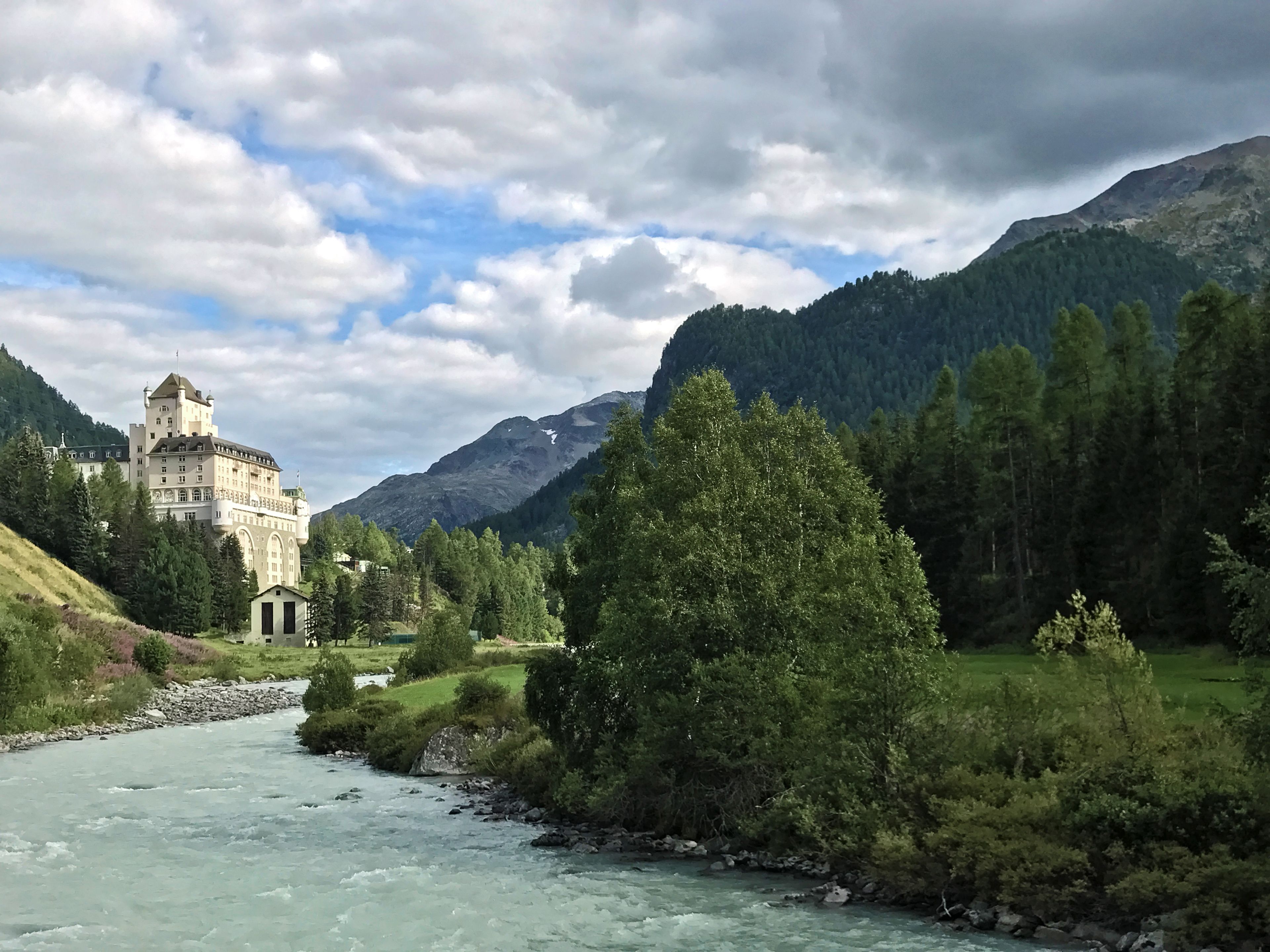Un bâtiment qui ressemble à un château, avec une rivière au premier plan et des montagnes en arrière-plan.