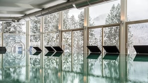 Una piscina interior con paredes de vidrio transparente que reflejan árboles nevados y montañas en el exterior.