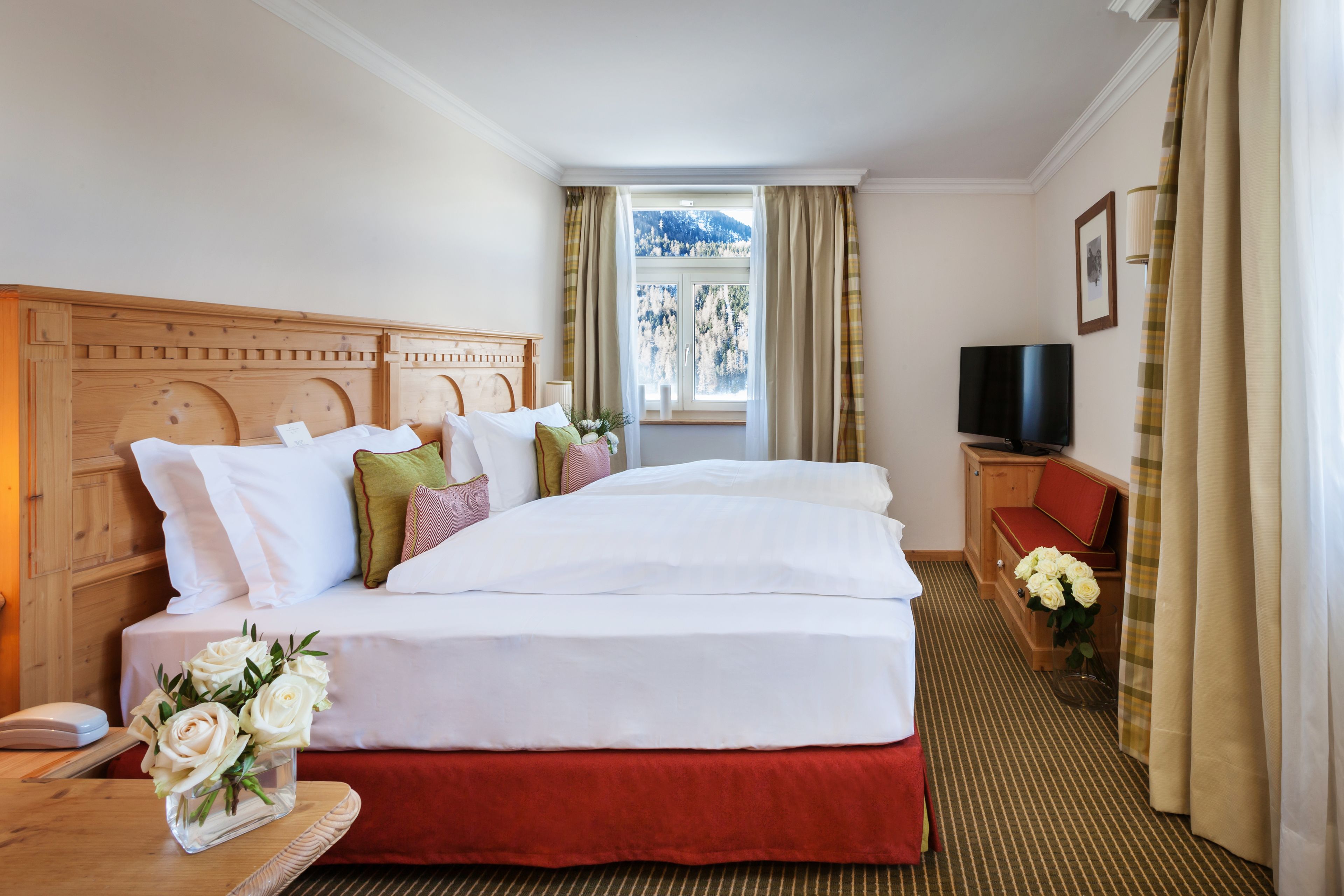 A hotel room with a large bed, a wooden headboard, a television, a desk with a telephone, and a window with curtains.