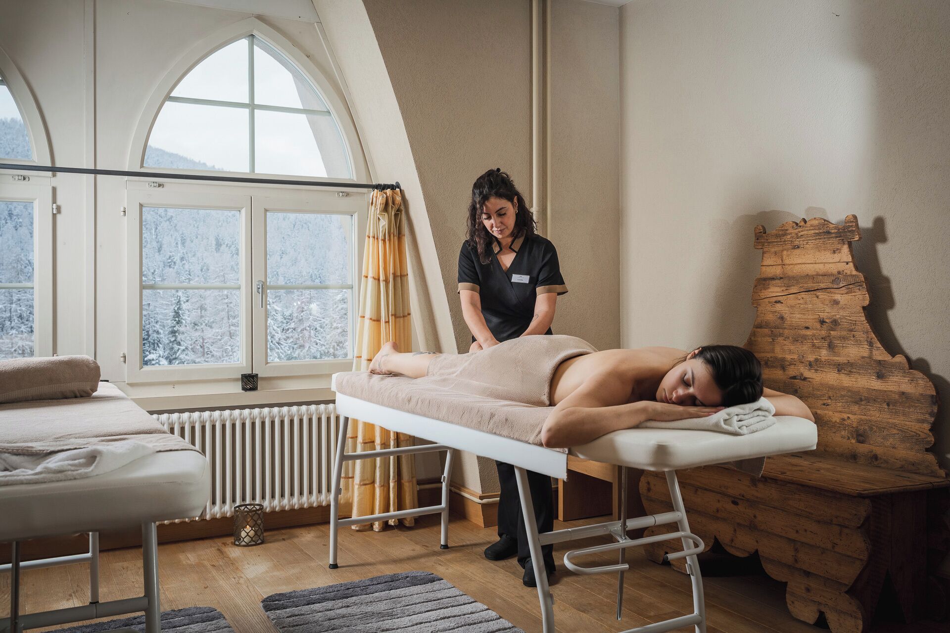 Ett rum för massagebehandlingar med en träpanel bakom massagebänken, en massagebänk och ett fönster med utsikt över snötäckta träd.