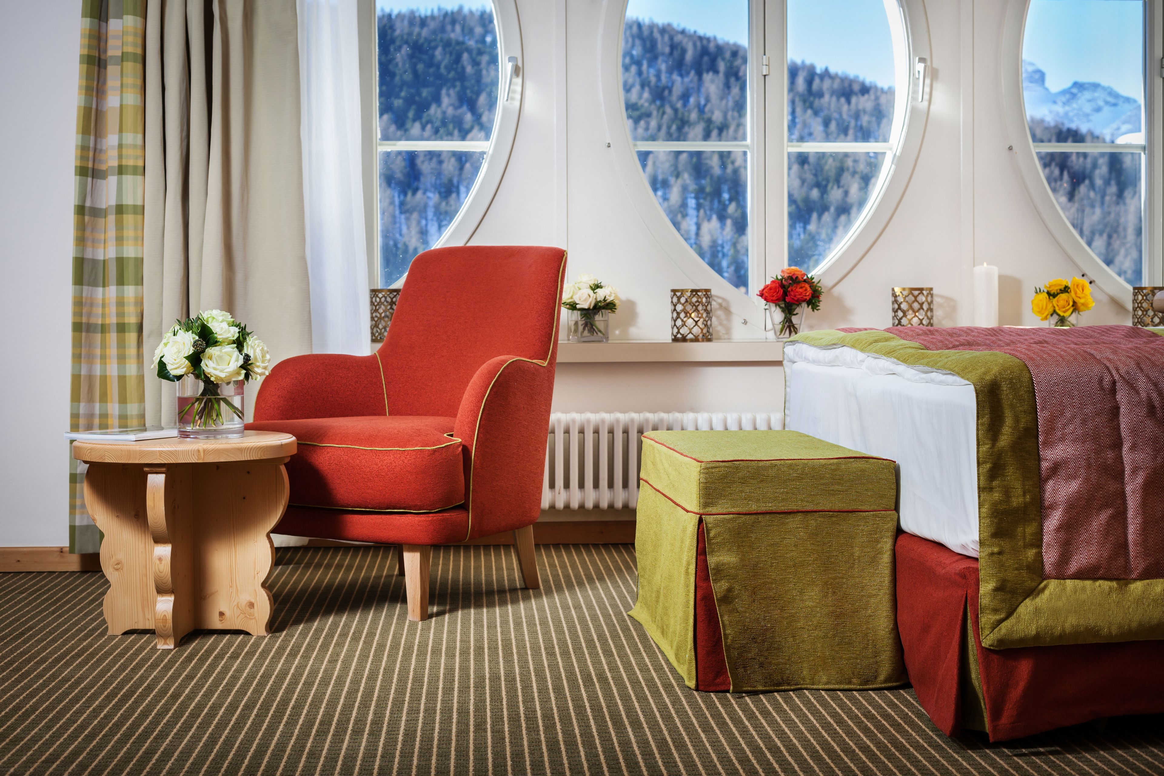 A room with a red chair, a small wooden table, a bed with a green and red bedspread, and a window with a view of snow-covered mountains.