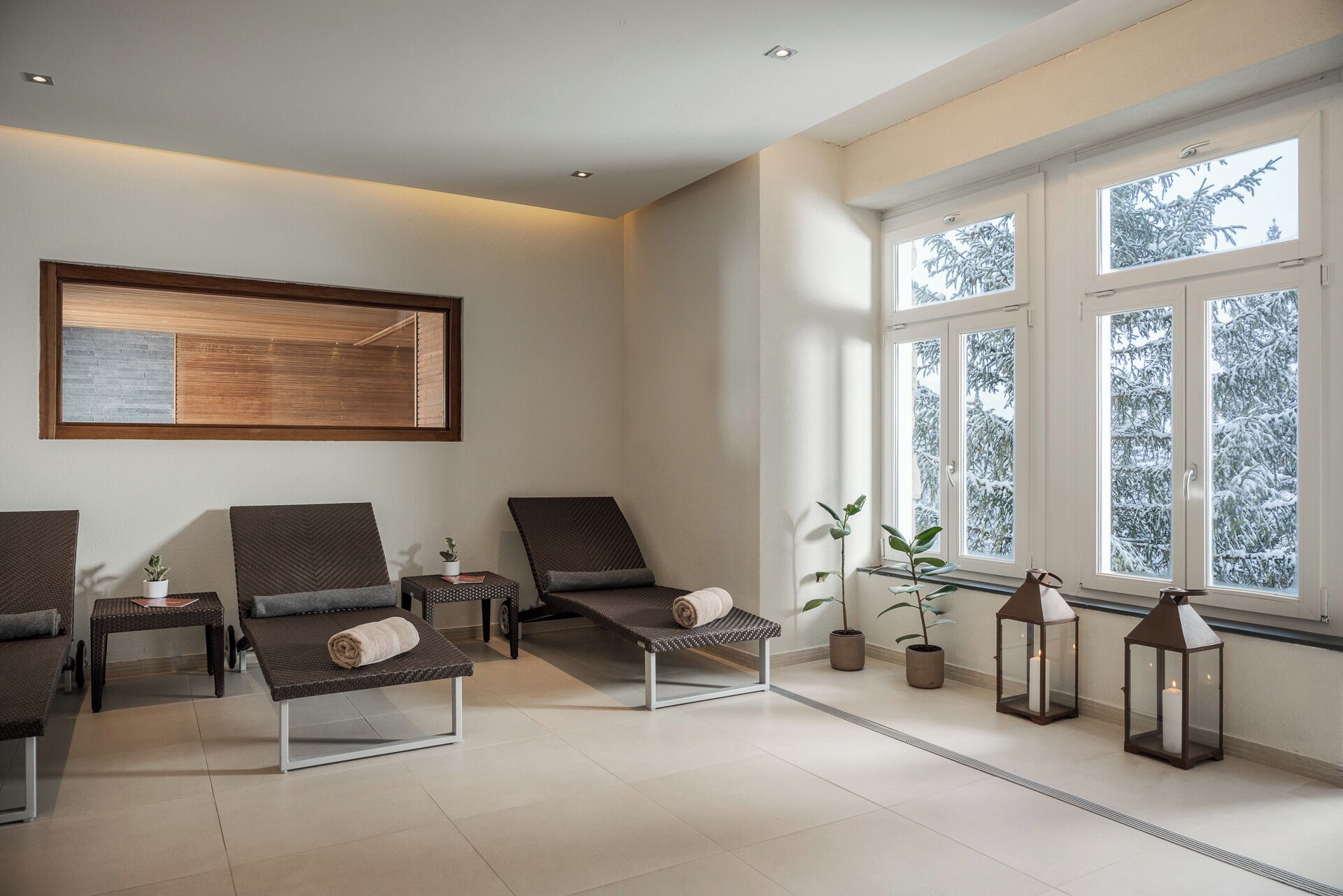 A modern lounge area with reclining chairs, a small table with a plant, and a window with a view of snow-covered trees.