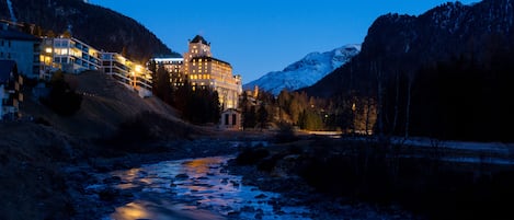 A hotel with a prominent building, a river in the foreground, and mountains in the background.